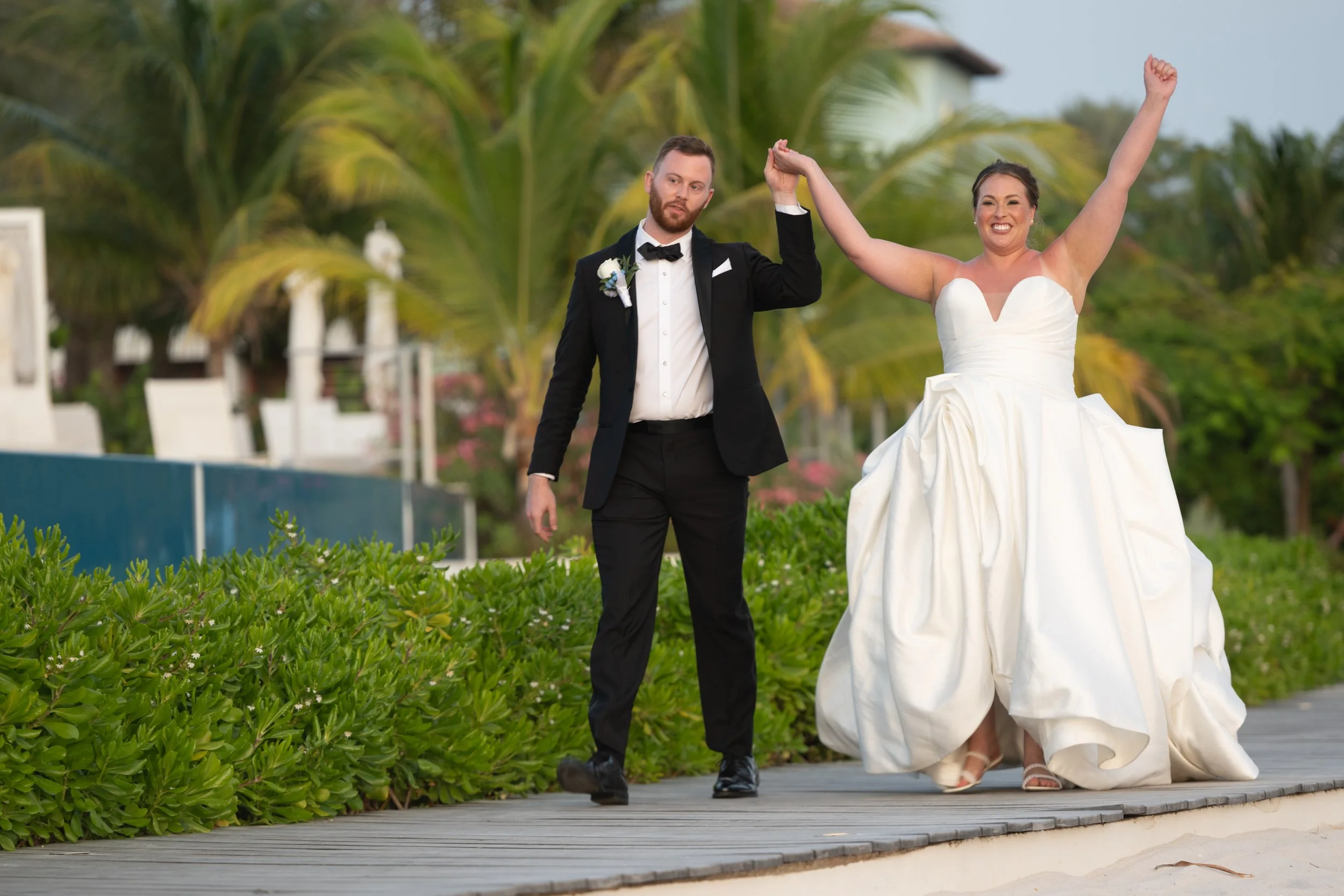 A newlywed couple, a man in a black tuxedo and a woman in a white wedding gown, walking hand in hand outdoors on a wooden pathway surrounded by greenery and palm trees.