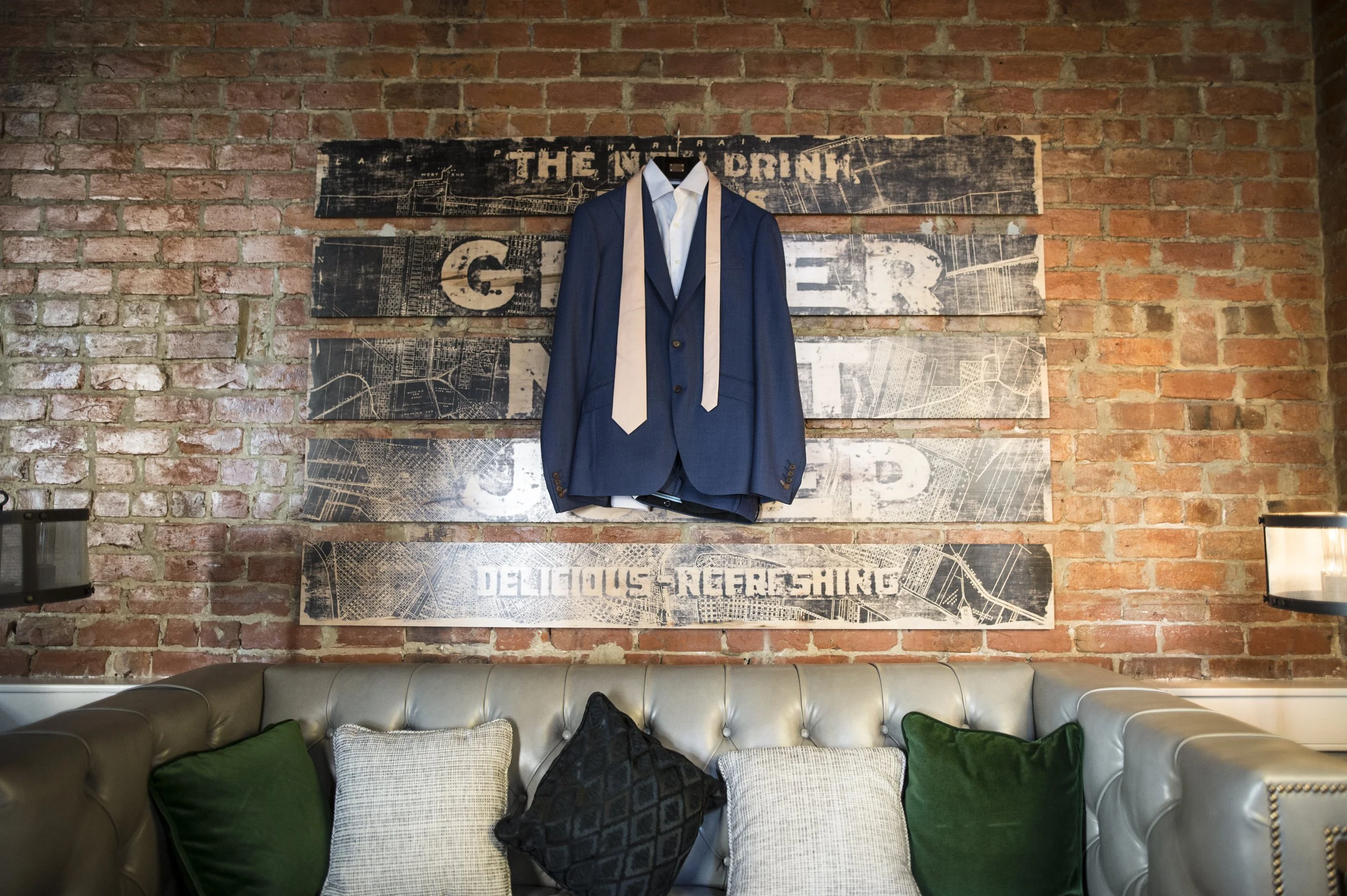 A brick wall with a distressed wooden sign hanging on it. The sign has parts of textual graphics, including the words 'THE' and 'GIR' visible. A navy blue suit jacket with a white shirt and beige tie is hanging on a hook, attached to the sign.