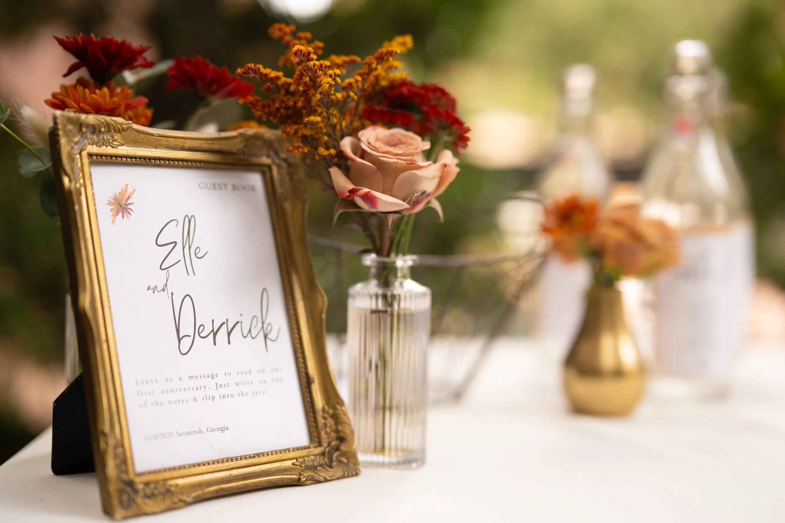 A decorative table setup featuring a gold picture frame with a guest book message, surrounded by colorful flowers in vases, including pink roses and red and orange blossoms, on a white table, with a blurred outdoor garden background.