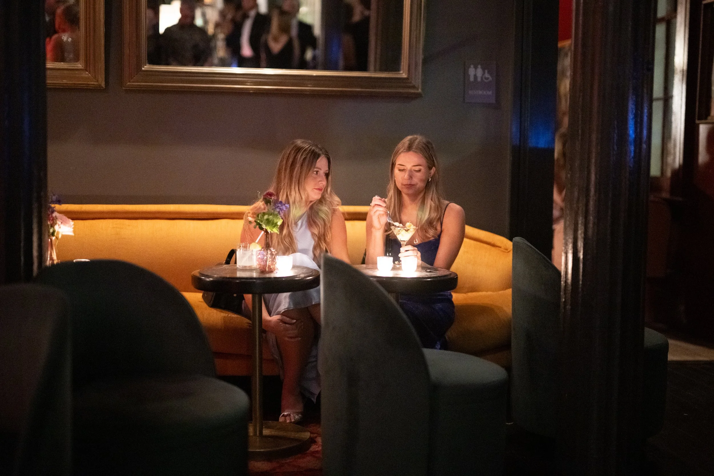 Two women sitting on a yellow couch at a darkened indoor restaurant or lounge, sharing a dessert. The background has framed artwork and a sign indicating restrooms. Candles and flowers on the table add to the ambiance.