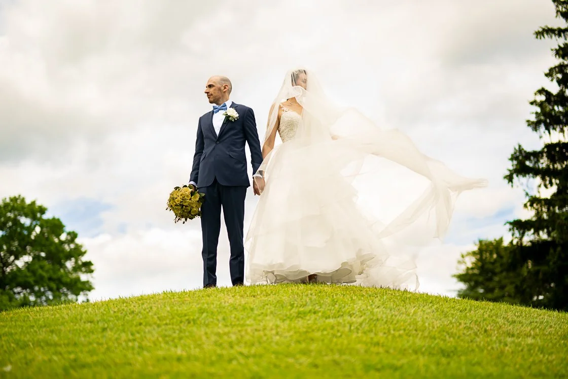 A bride and groom holding hands outdoors on a grassy hill, with the bride in a white wedding gown and veil, and the groom in a navy suit holding a bouquet of flowers.