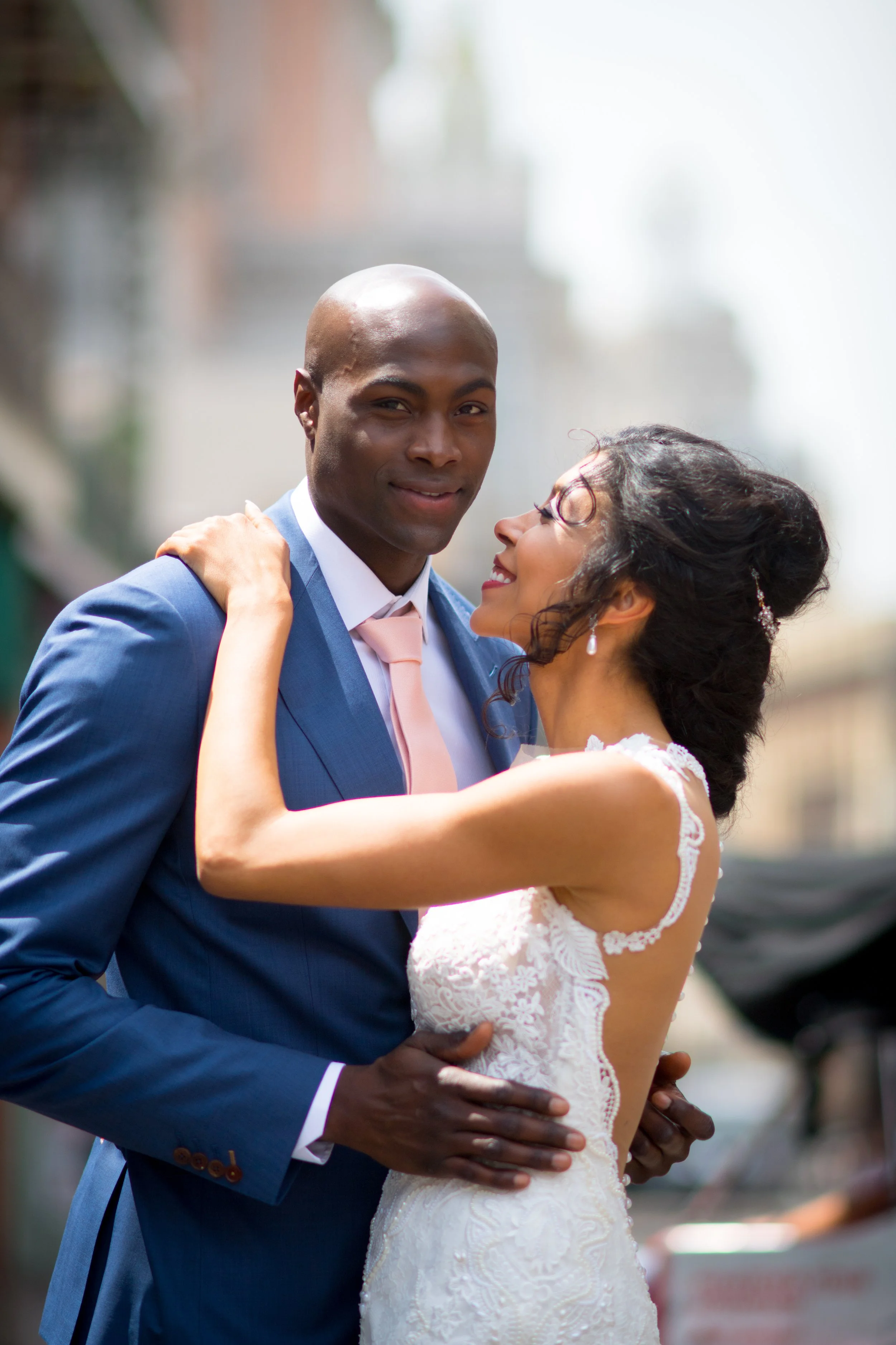 A newlywed couple dancing outdoors, with the bride in a white lace dress and the groom in a blue suit and pink tie, sharing a tender moment.