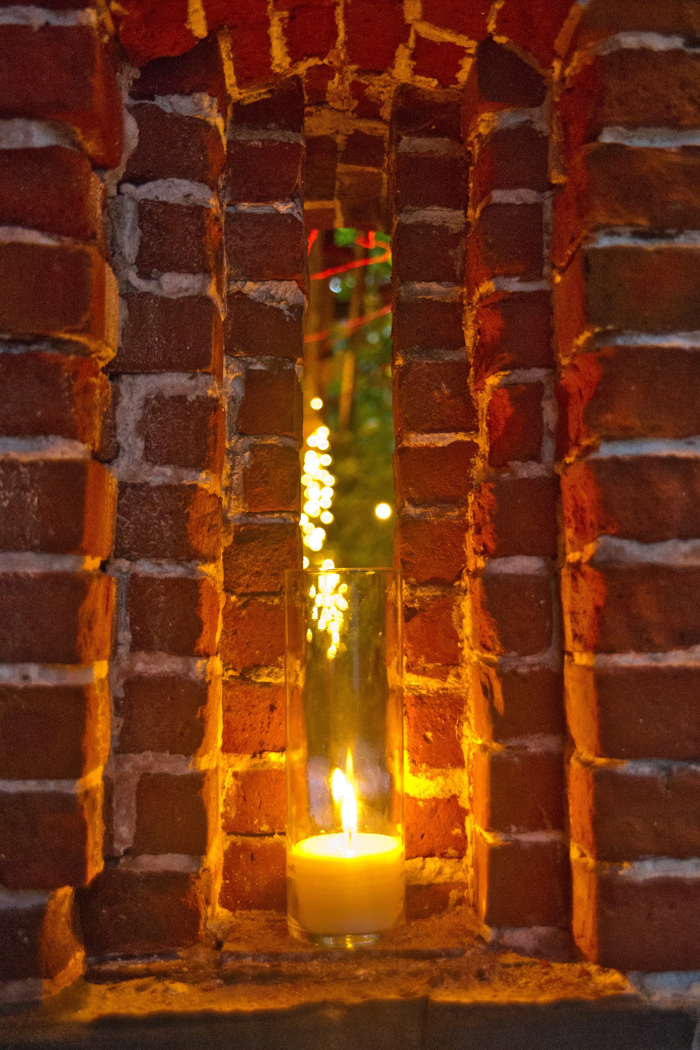 A candle in a glass holder placed on a brick wall ledge with a small opening revealing blurred colorful lights in the background.