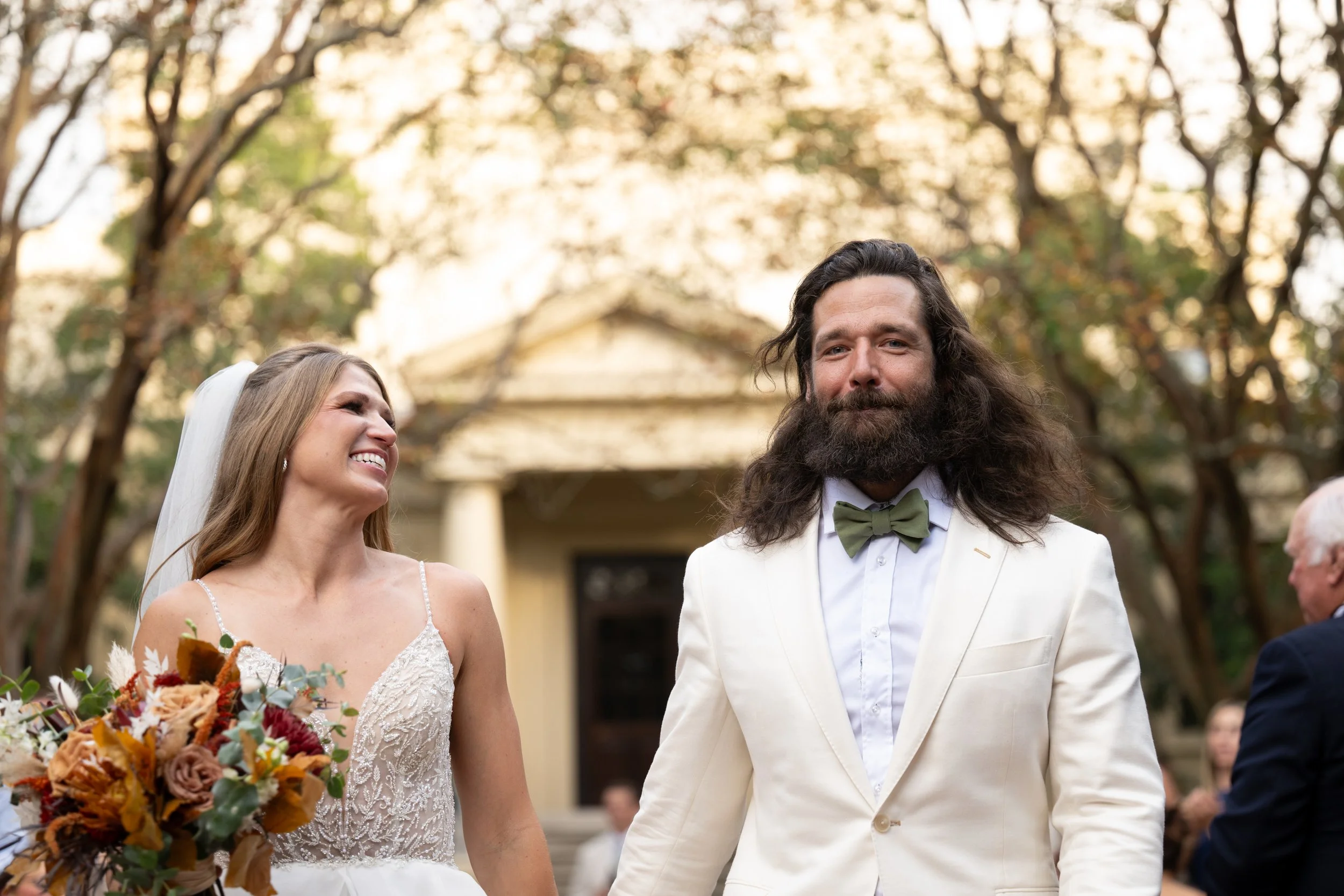 A bride and groom at their outdoor wedding, with the bride holding a bouquet of colorful flowers, smiling and looking at each other, surrounded by trees with autumn leaves.