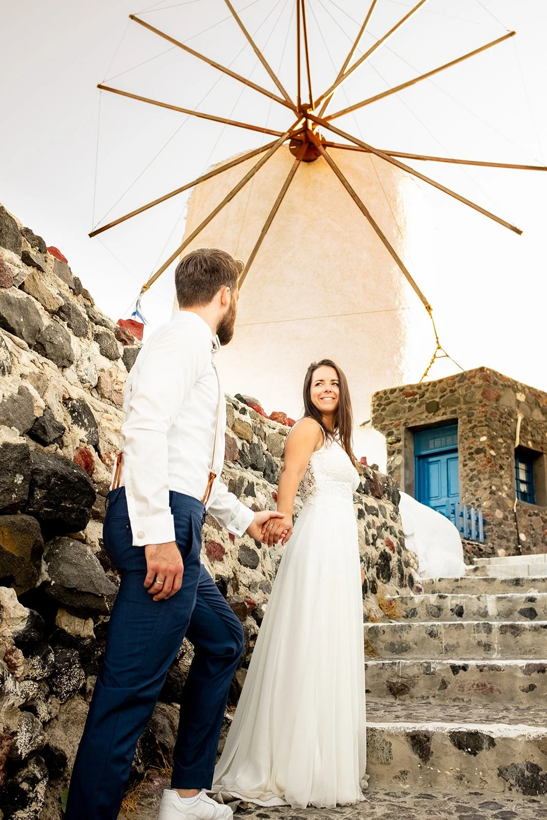 A couple, a man and a woman, holding hands and standing on stone steps outdoors under a large cream-colored parasol, with a small stone building with a blue door in the background.