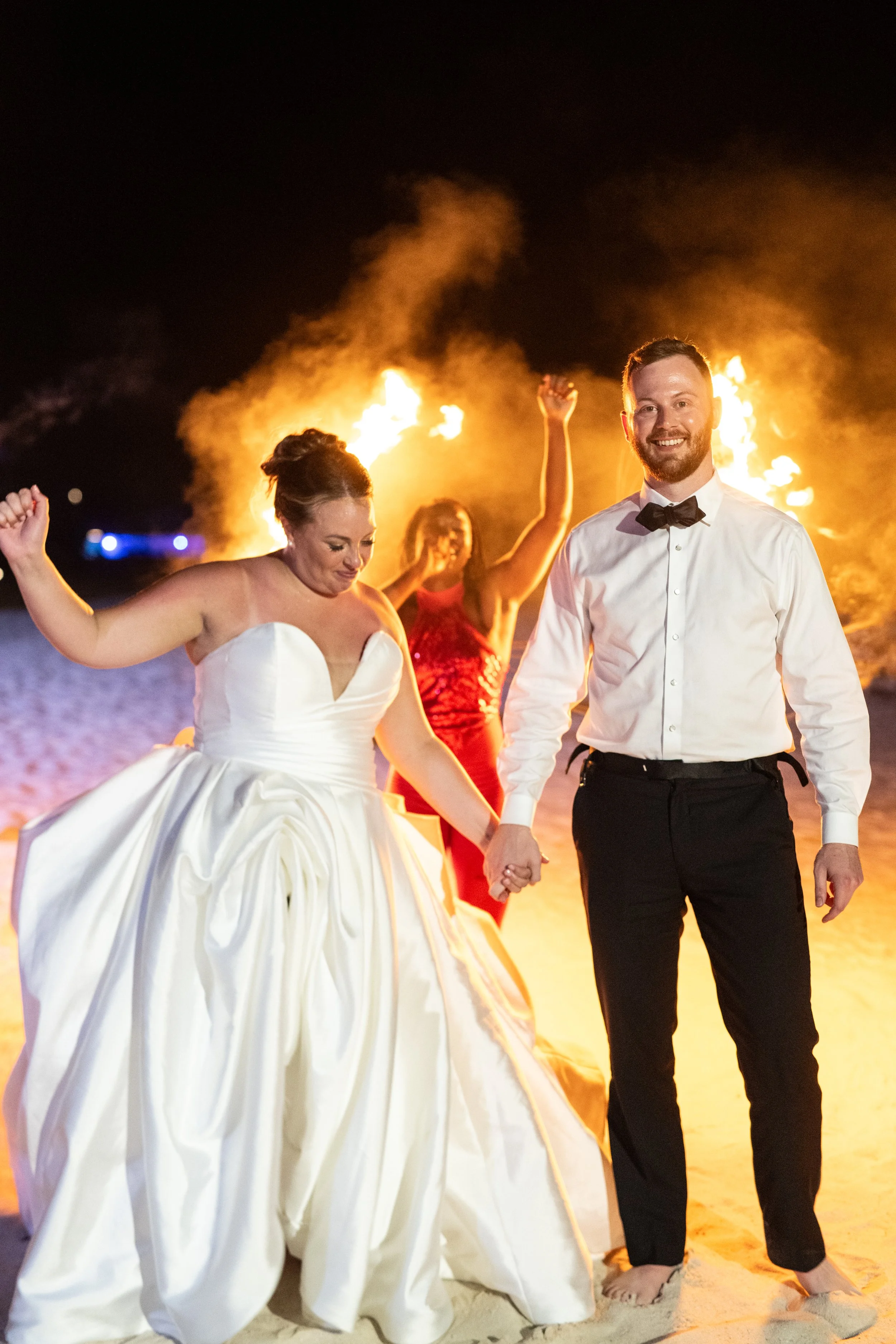 A smiling man in a white shirt and black bow tie holding hands with a woman in a strapless white wedding dress walking on the beach at night. Two women with fire torches are dancing in the background.