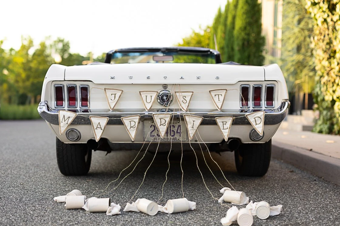 The back of a white vintage Ford Mustang with a 'Just Married' banner made of paper triangles hanging from the trunk. The banner letters spell out 'JUST MARRIED.' Tin cans attached to the bumper with strings are scattered on the ground behind the car