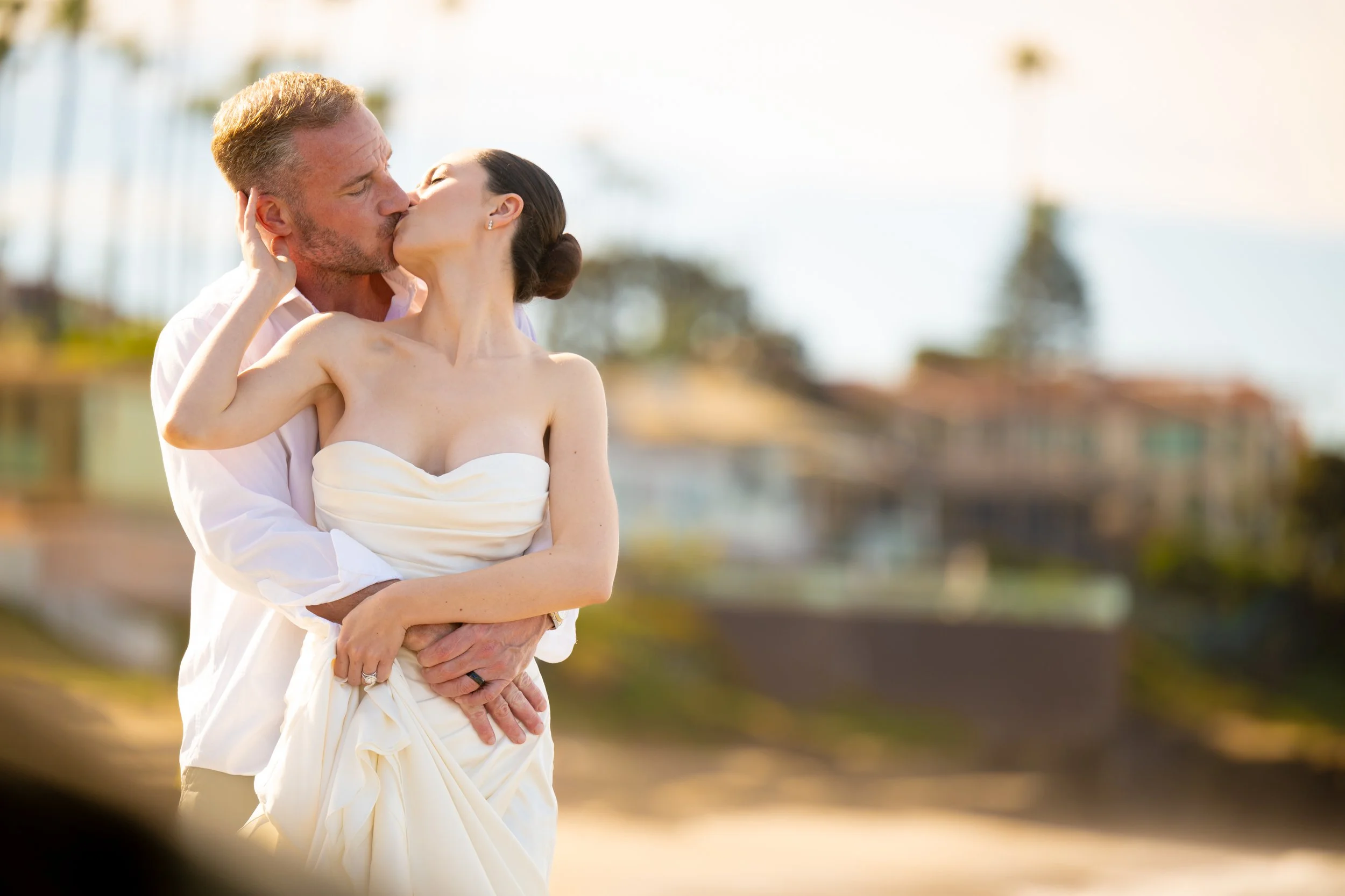 A couple sharing a romantic kiss outdoors. The man is holding the woman in his arms, and she is wearing a strapless white dress. The background shows a blurred outdoor setting with trees and a bright sky.