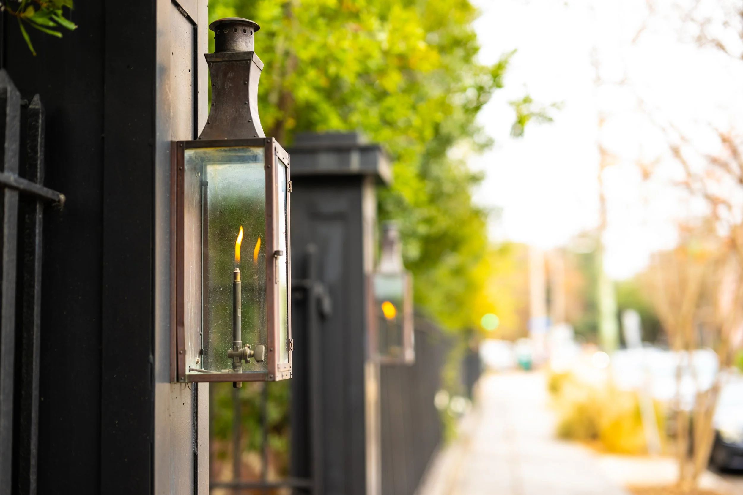 A metal outdoor lantern with a glass front, mounted on a black wall, with trees and sunlight in the background.