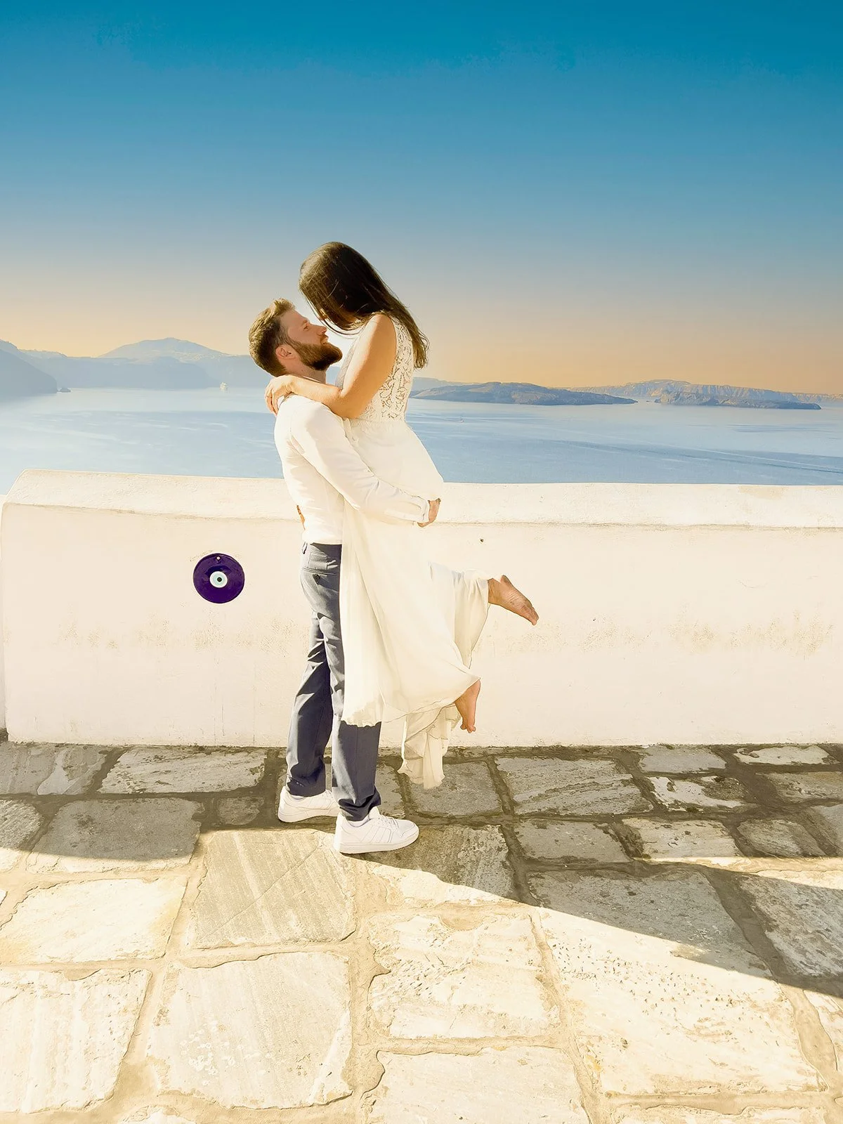 A man lifts a woman in a white dress against a backdrop of a large body of water, mountains, and a clear blue sky during sunset, with a white wall and stone pavement in the foreground.