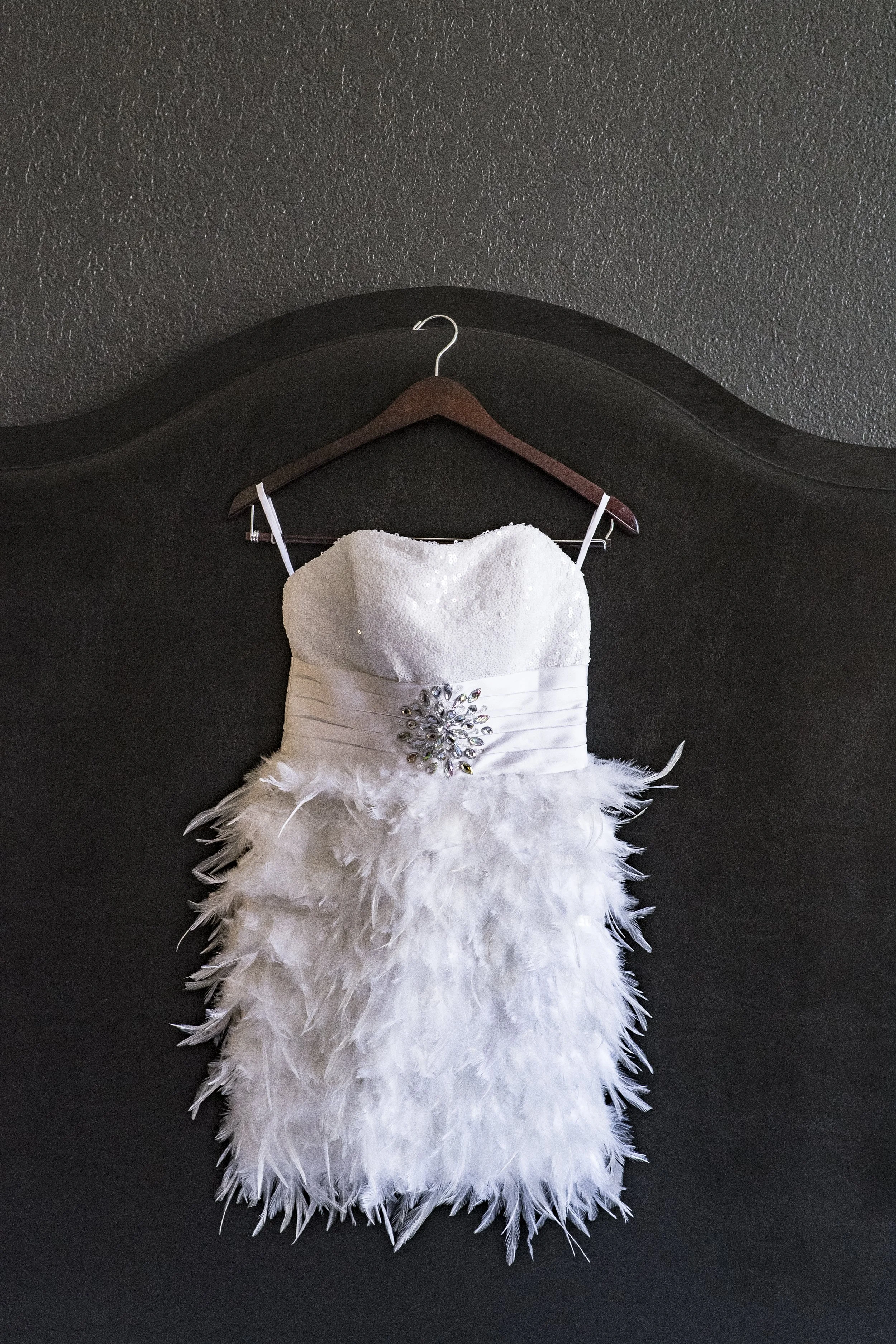A white dress with feather details and a jeweled brooch on the bodice, hanging on a wooden hanger against a dark wall.