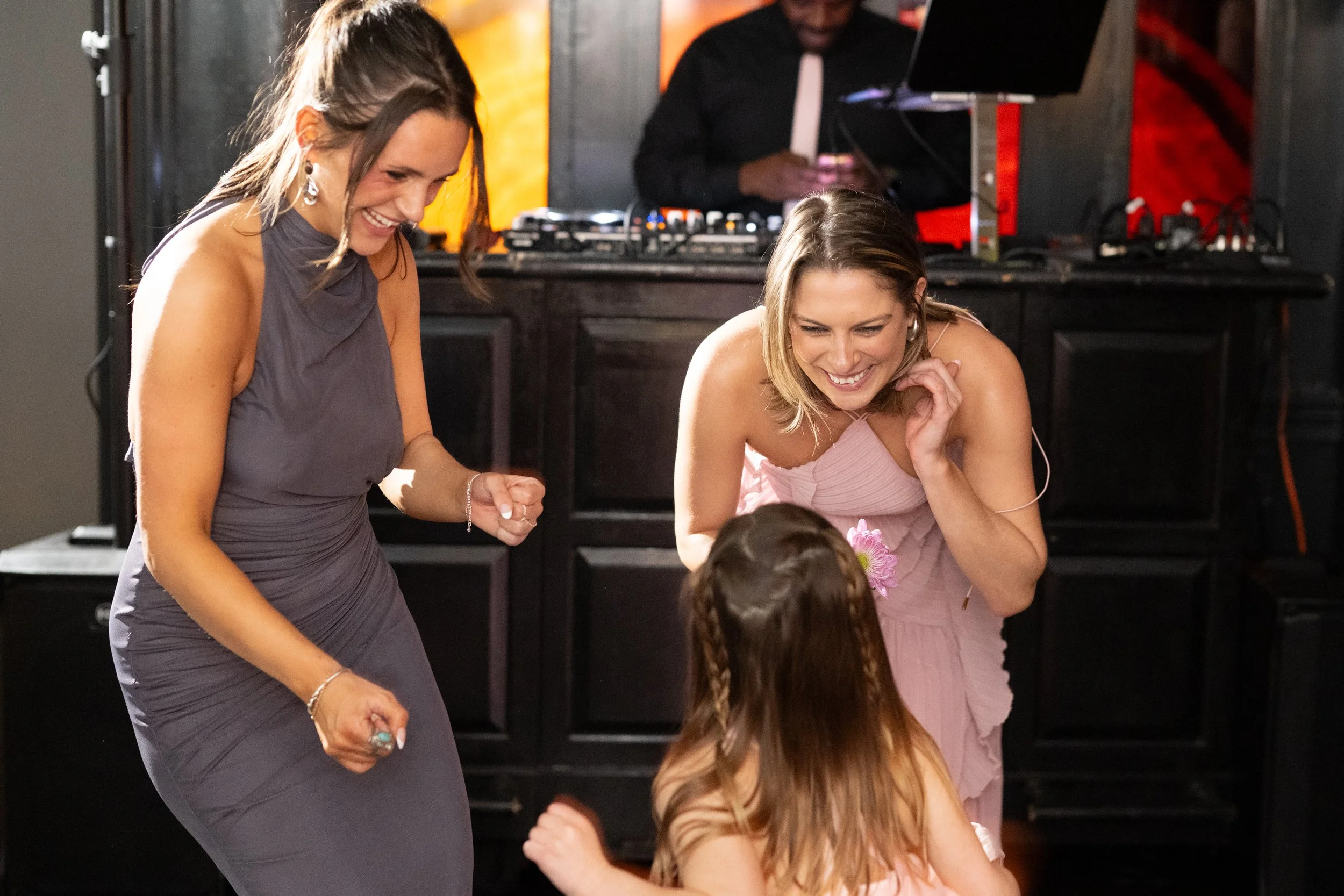 Two women smiling and interacting with a child at a social event, with a DJ setup and a laptop in the background.