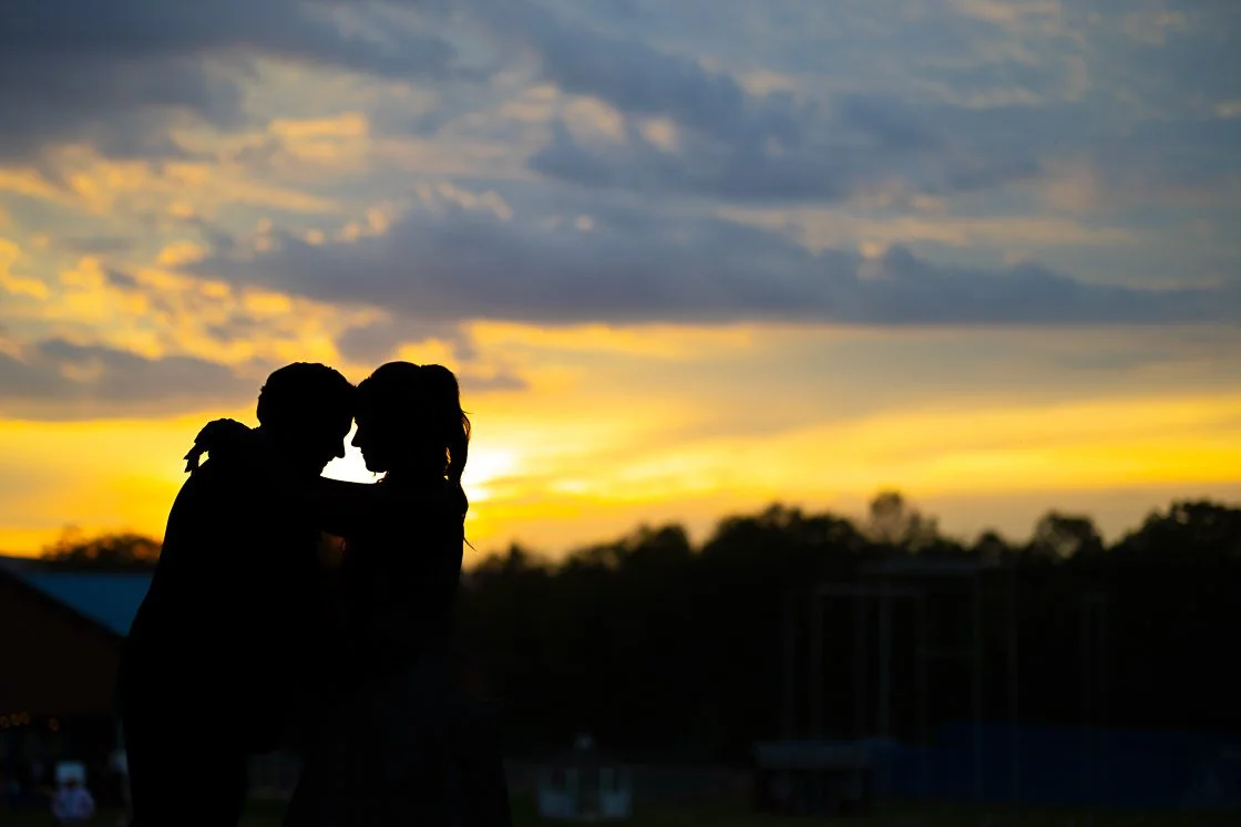 Silhouettes of a couple kissing against a sunset sky with clouds and trees in the background.