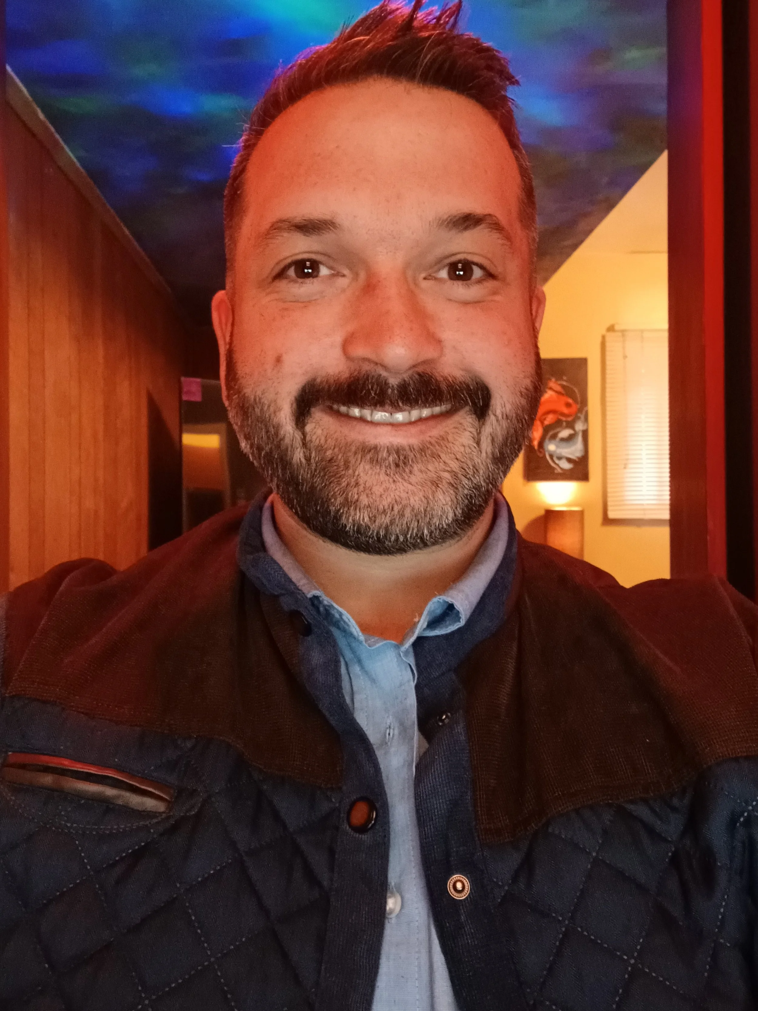 A smiling man with short brown hair, a beard, and wearing a blue shirt and dark quilted jacket indoors with colorful lighting and artwork on the wall.