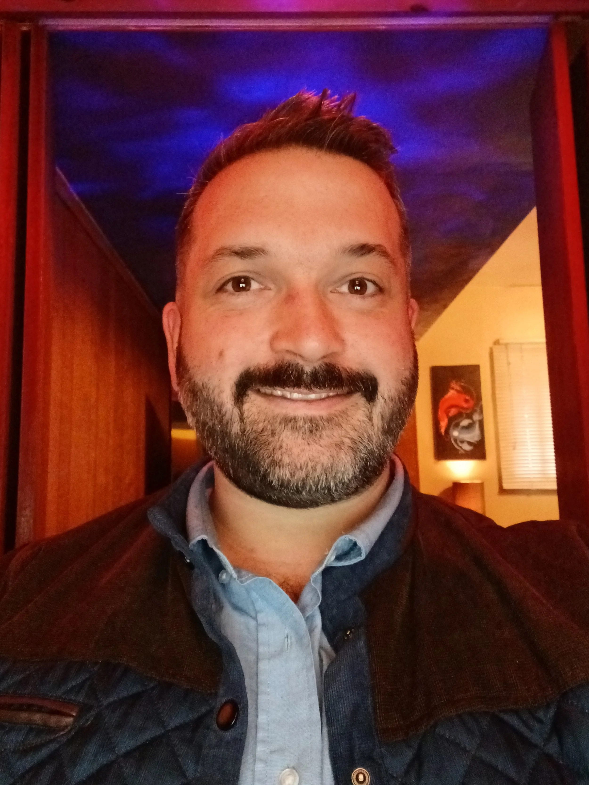 A smiling man with short brown hair, a beard, and mustache, wearing a light blue collared shirt and a brown jacket, takes a selfie indoors with colorful lighting and artwork in the background.