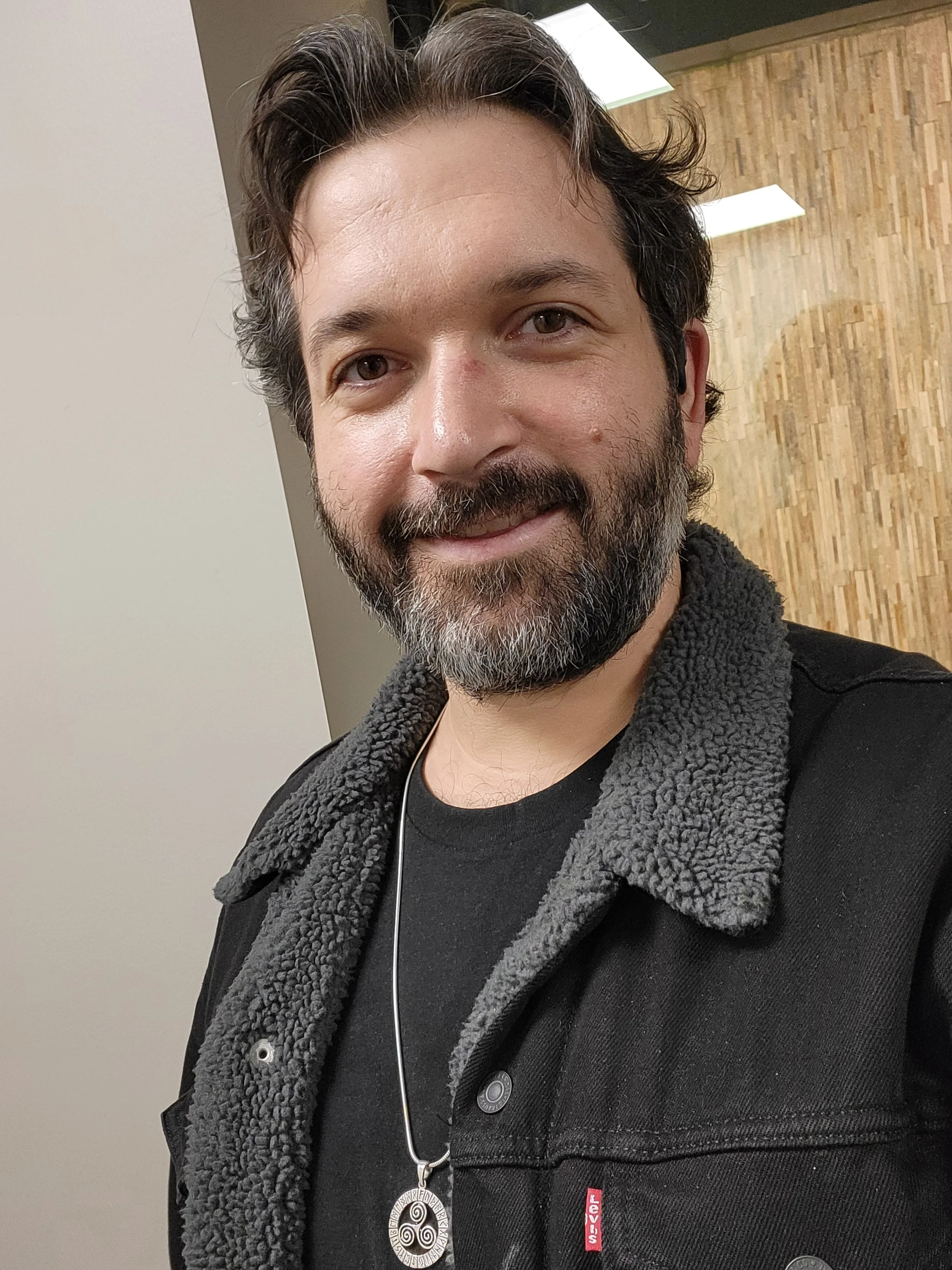 A man with dark hair and a beard smiling for a selfie indoors, wearing a black shirt, a black jacket with a sherpa lining, and a necklace with a circular pendant.