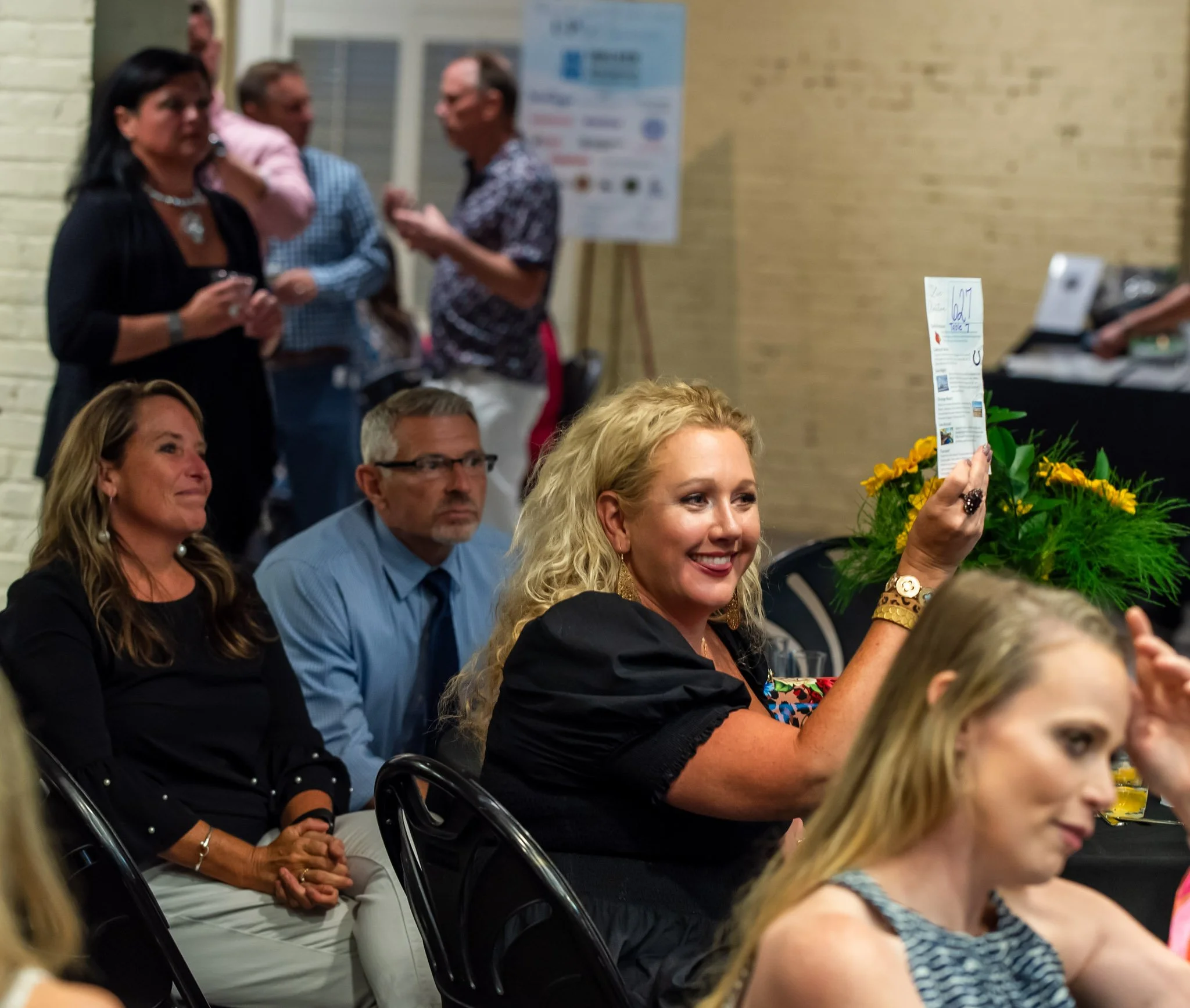 A woman with blonde curly hair smiling and holding up a piece of paper at a seated event, with other seated and standing people in the background.