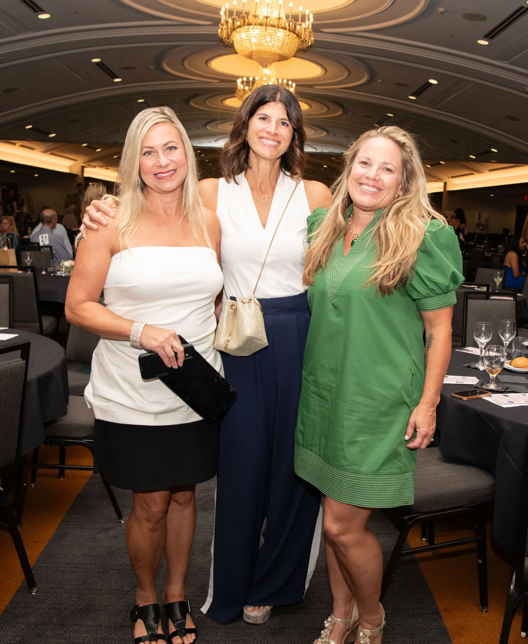 Three women smiling and posing at a formal event or dinner in a banquet hall with chandeliers.