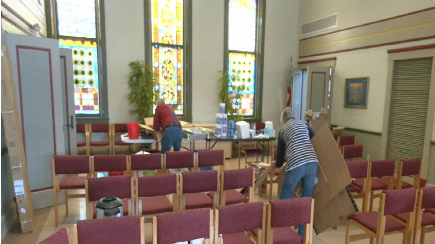 Two people are setting up or cleaning a church or meeting room with rows of red-upholstered chairs. There are large, colorful stained glass windows in the background and tables with cleaning supplies and boxes.