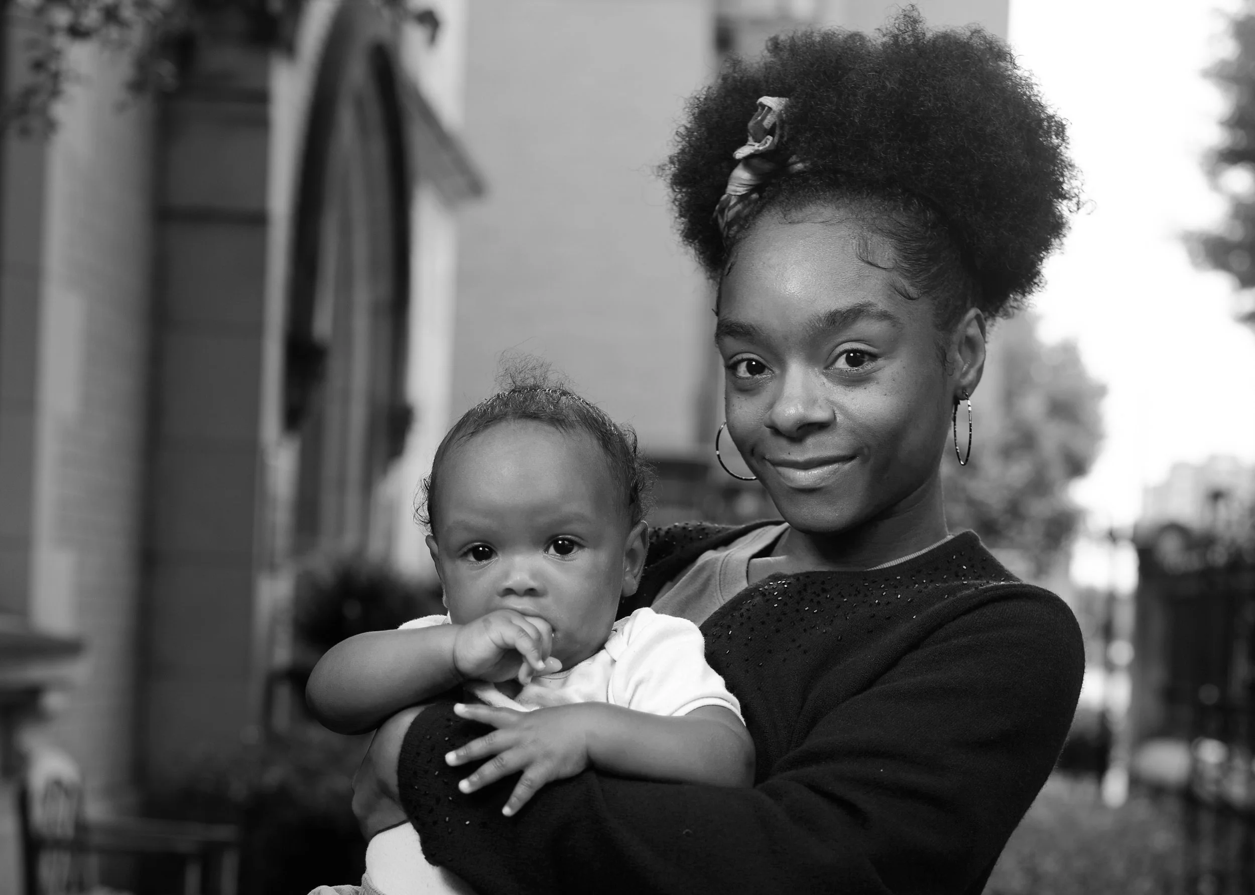 A woman holding a baby outdoors, both looking at the camera, in black and white.