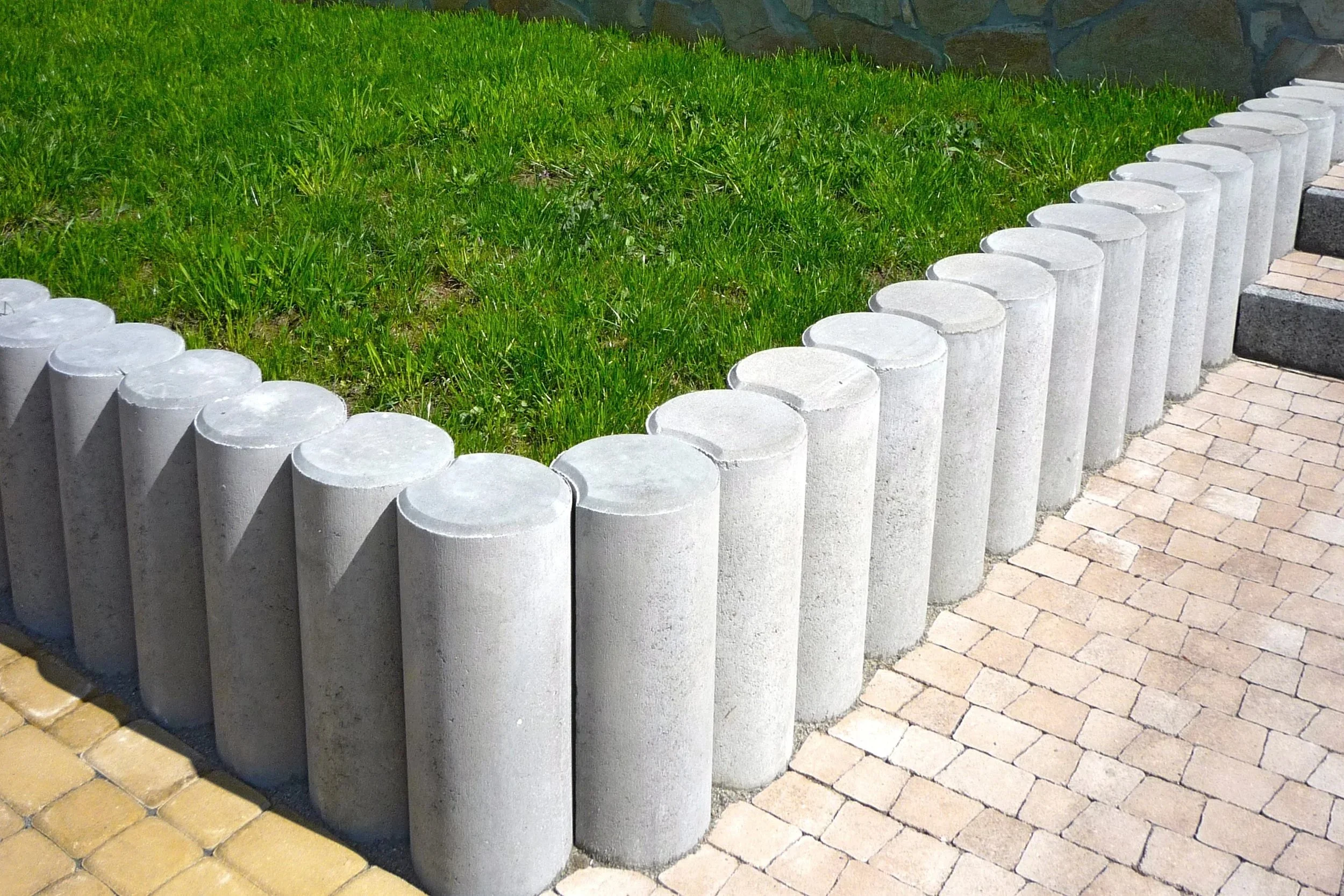 Concrete bollards forming a curved border alongside a grassy area and a stone-paved walkway.