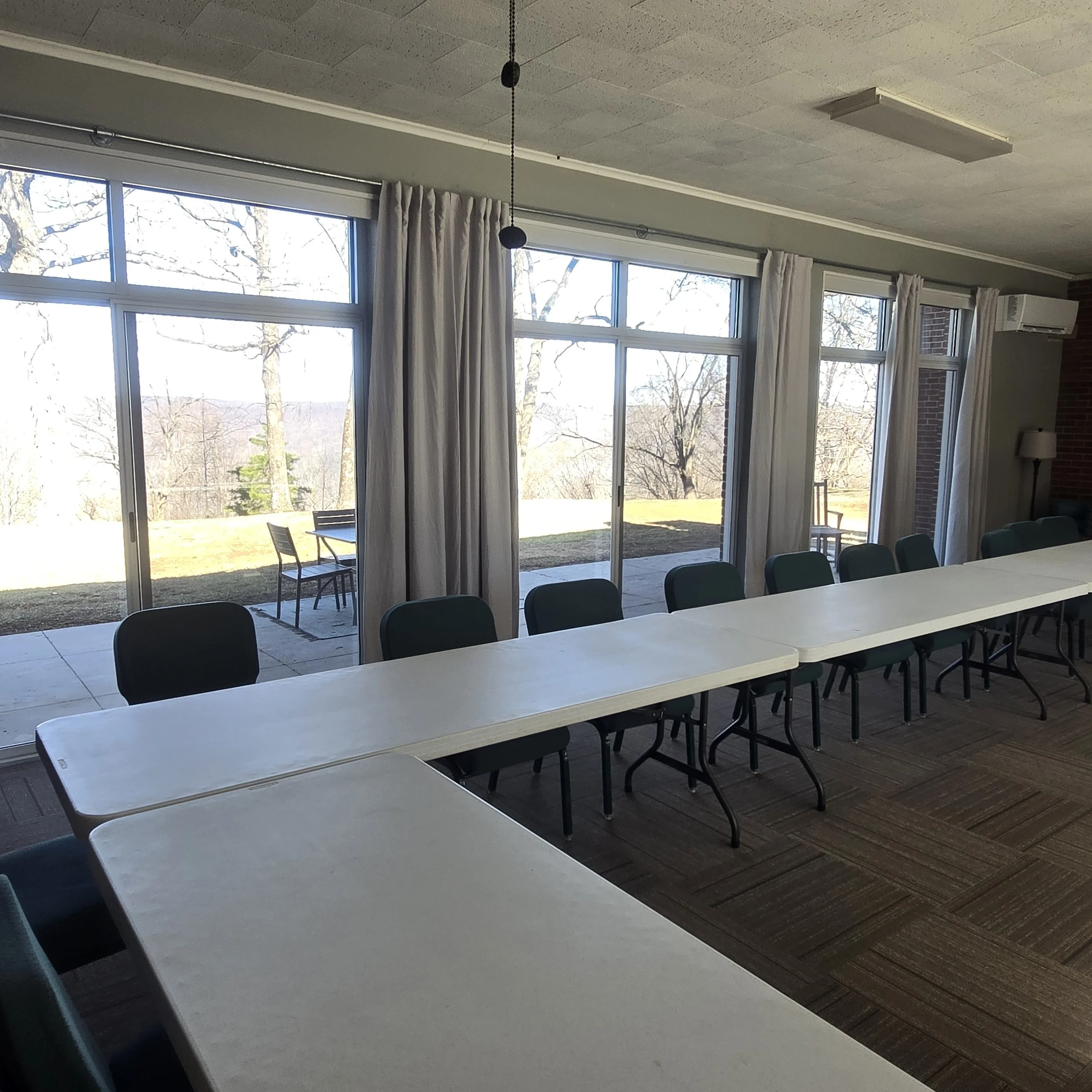 Indoor meeting room with white rectangular tables and green chairs arranged along the tables, large glass sliding doors with white curtains showing an outdoor patio with trees and chairs, carpeted floor, ceiling light fixtures, and air conditioning u