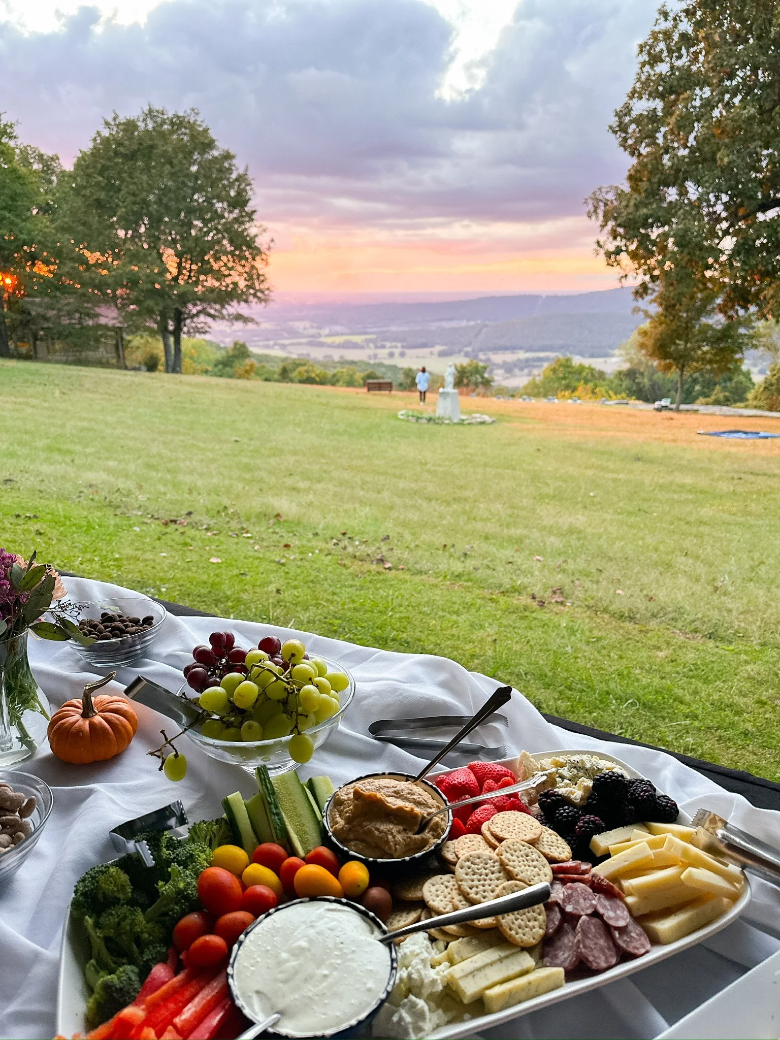 A picnic table with various foods including grapes, cherry tomatoes, cheese, crackers, vegetables, dips, and a small pumpkin, set outdoors with a scenic view of rolling hills and a sunset sky in the background.