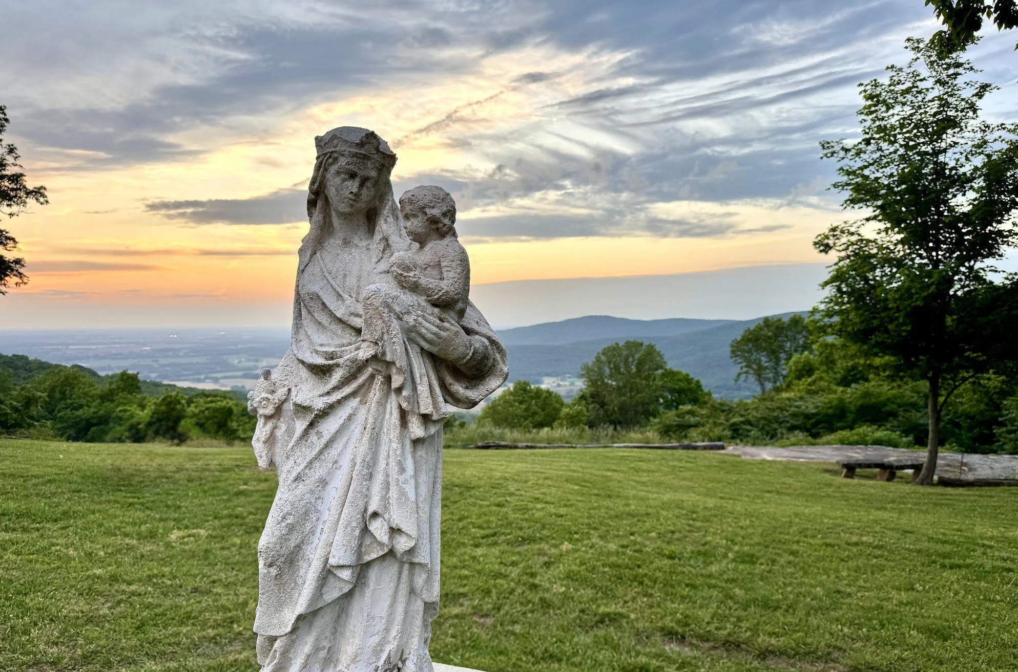 A stone statue of a woman holding a child, set outdoors on a grassy area at sunset, with hills, trees, and a partly cloudy sky in the background.