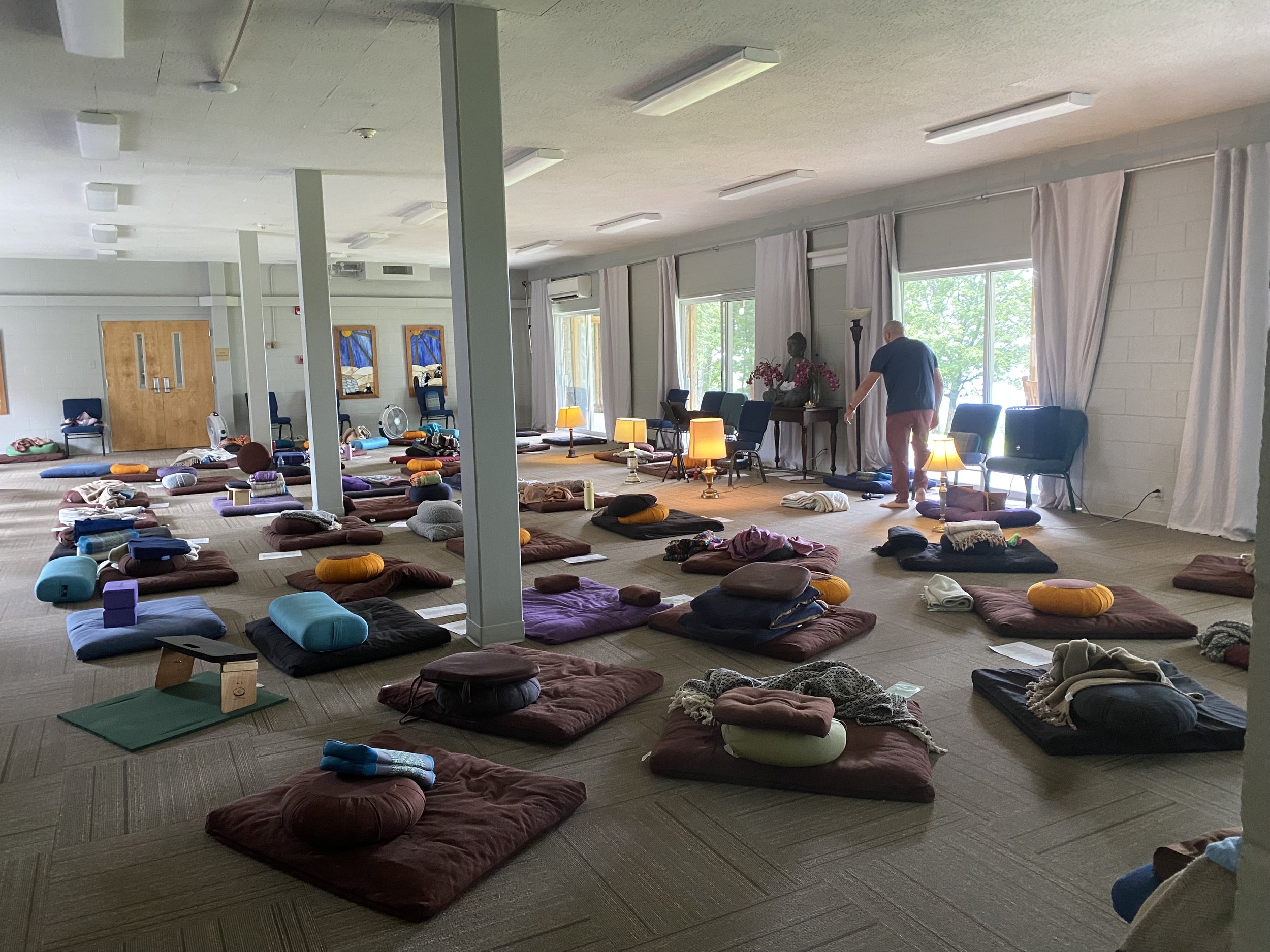 Indoor meditation space with cushions, bolsters, and blankets arranged on the floor. There are a few lamps and chairs near large windows with curtains, and a person standing near the windows.