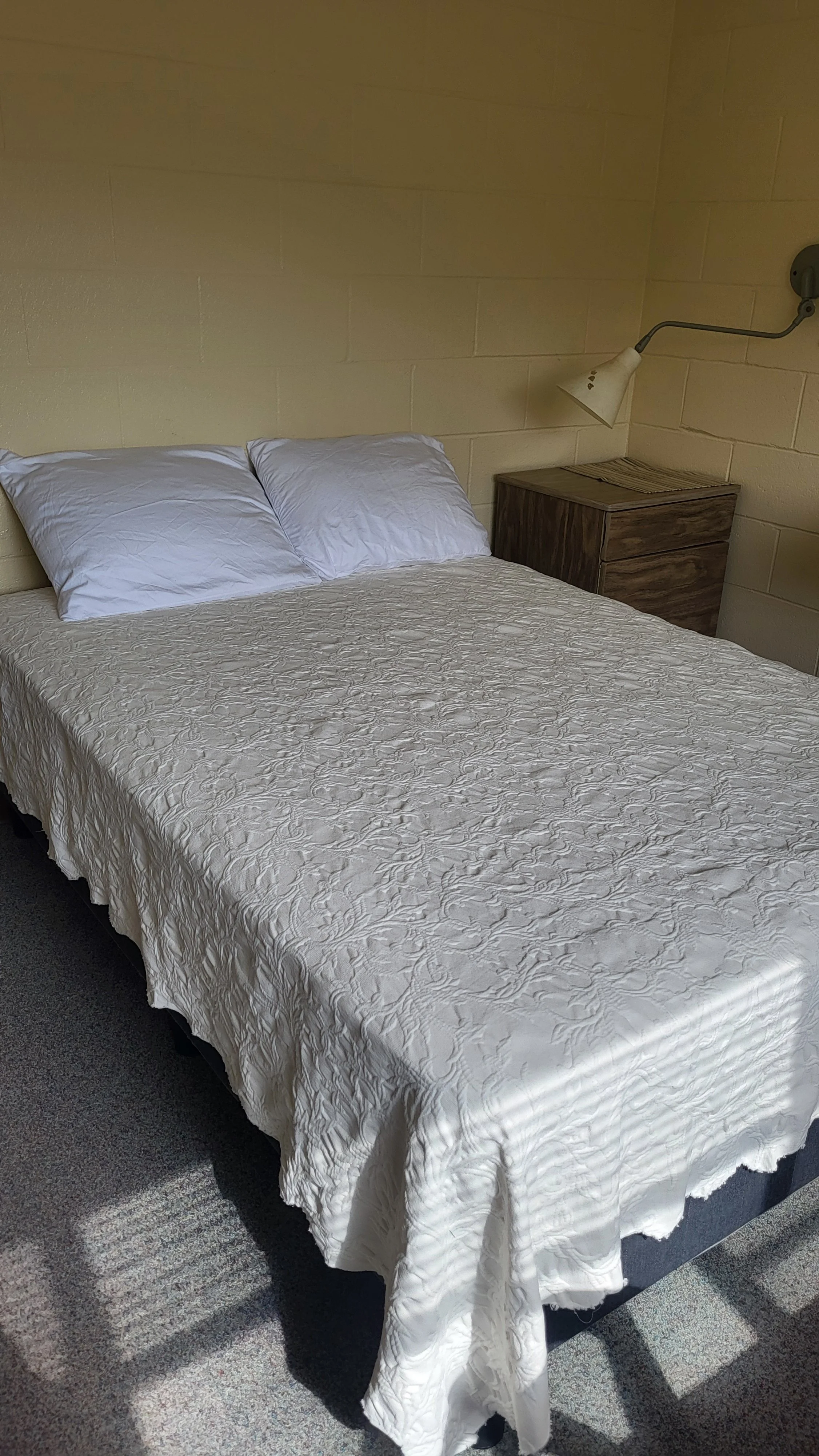 A bed with a white textured bedspread and two white pillows, next to a wooden nightstand, with a wall-mounted reading lamp on a beige brick wall.