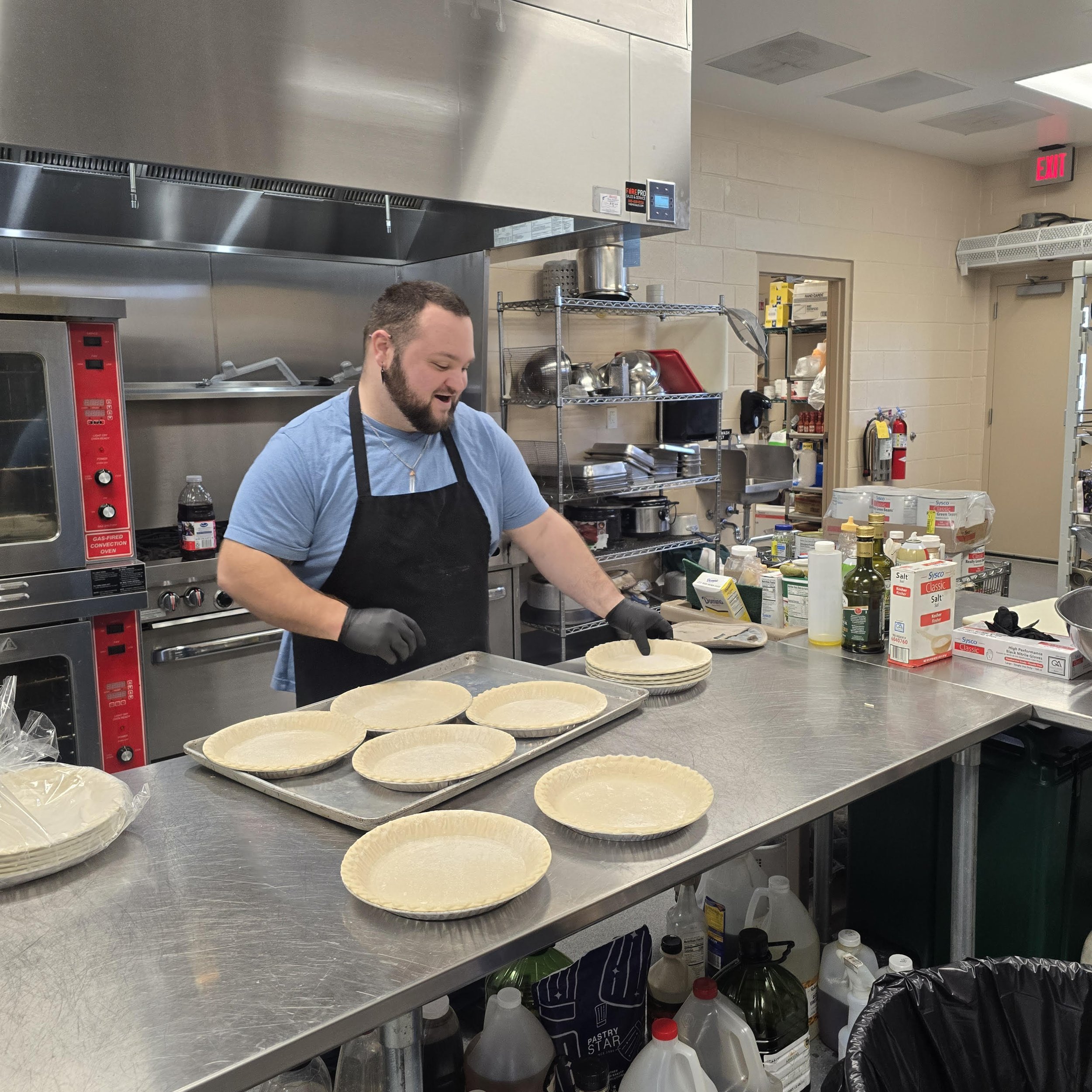 A man wearing a black apron and gloves preparing pie crusts in a commercial kitchen.