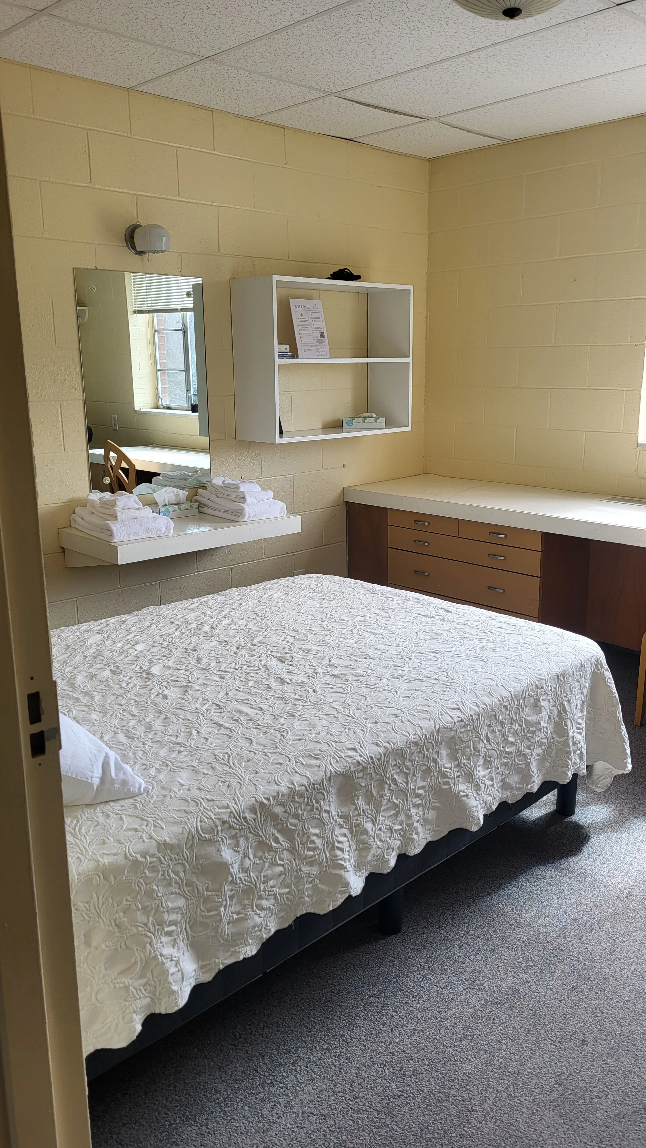 A simple hotel room with a bed covered in a white textured bedspread, a small mirror, folded towels, and a window.