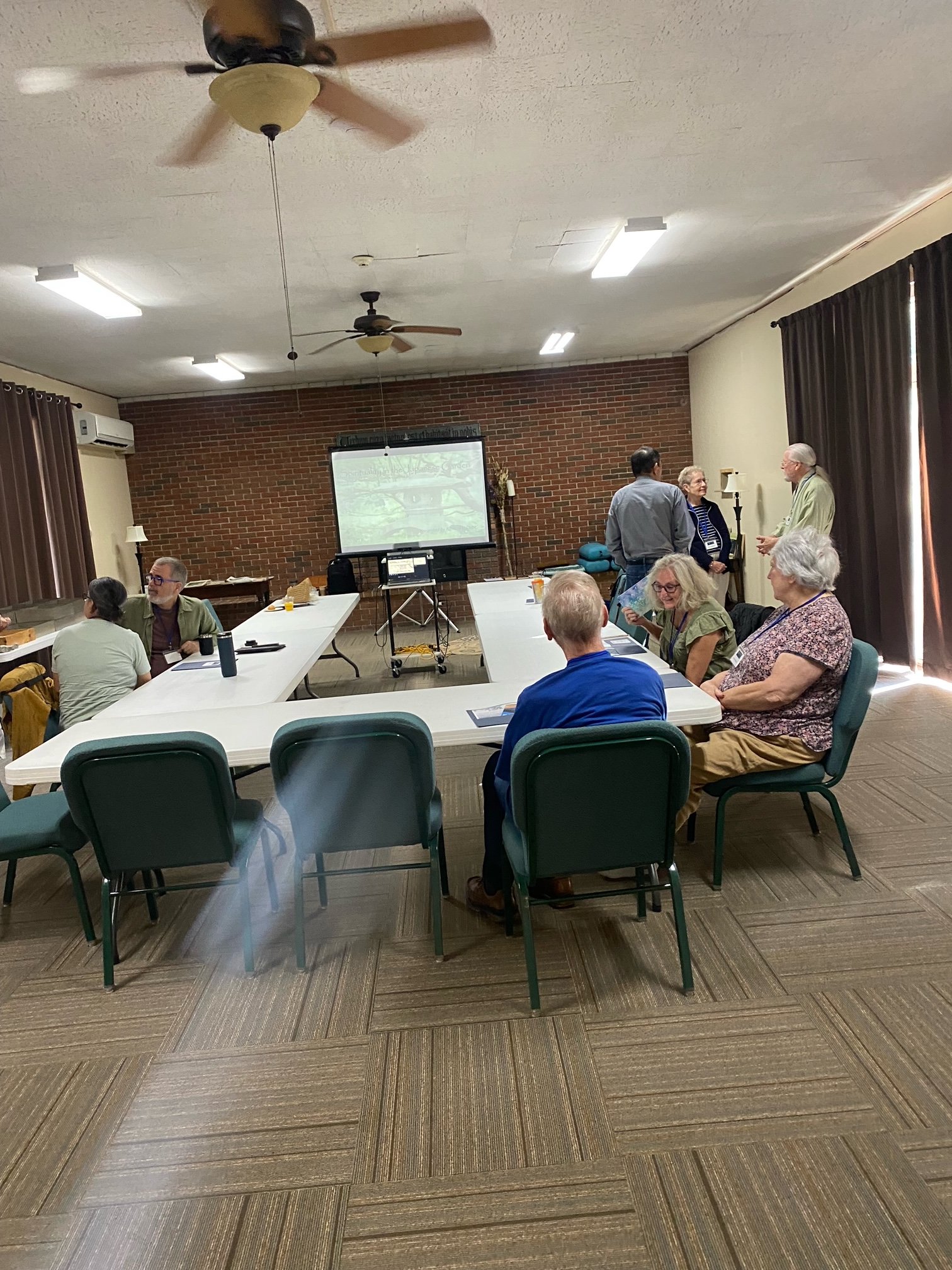 A group of elderly people gathered in a room around tables, some sitting and some standing, engaging in conversation. There is a projector screen at the front of the room displaying an image, and the room has brown curtains and a brick wall at the ba