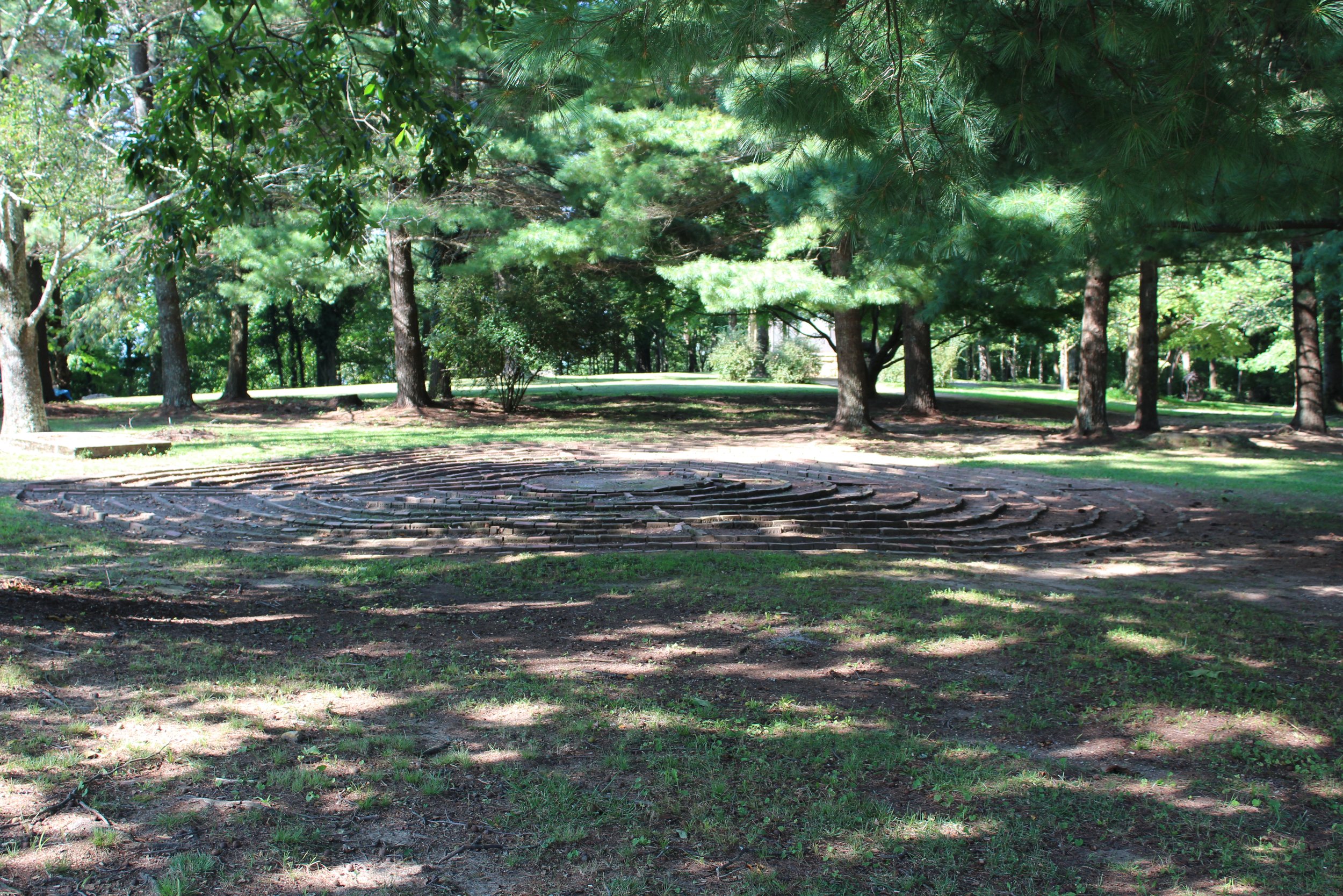 A park scene with a circular spiral brick sculpture on the ground, surrounded by green grass and tall trees with dense foliage, casting shadows on the ground.