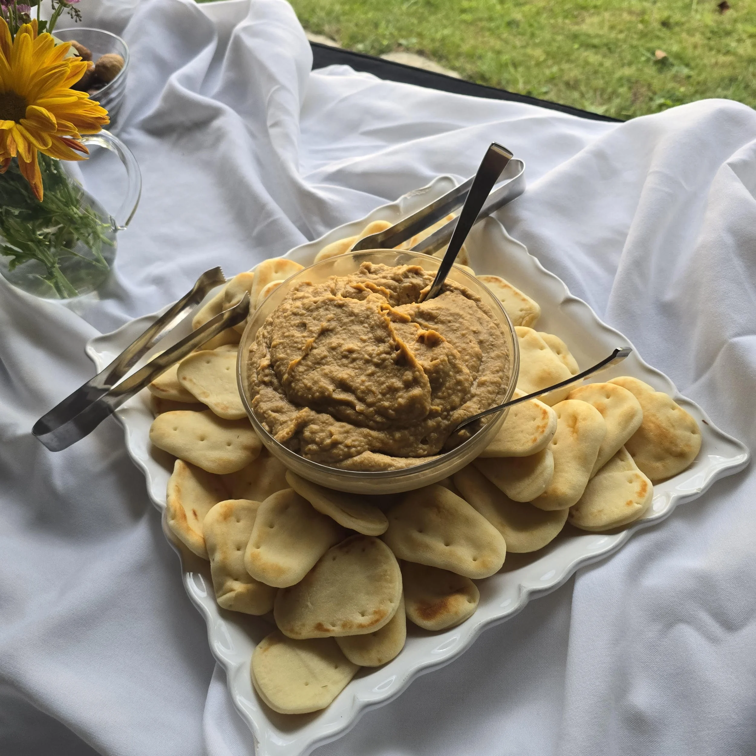 A serving plate with pita bread and a bowl of hummus, set on a white cloth with a flower bouquet nearby.