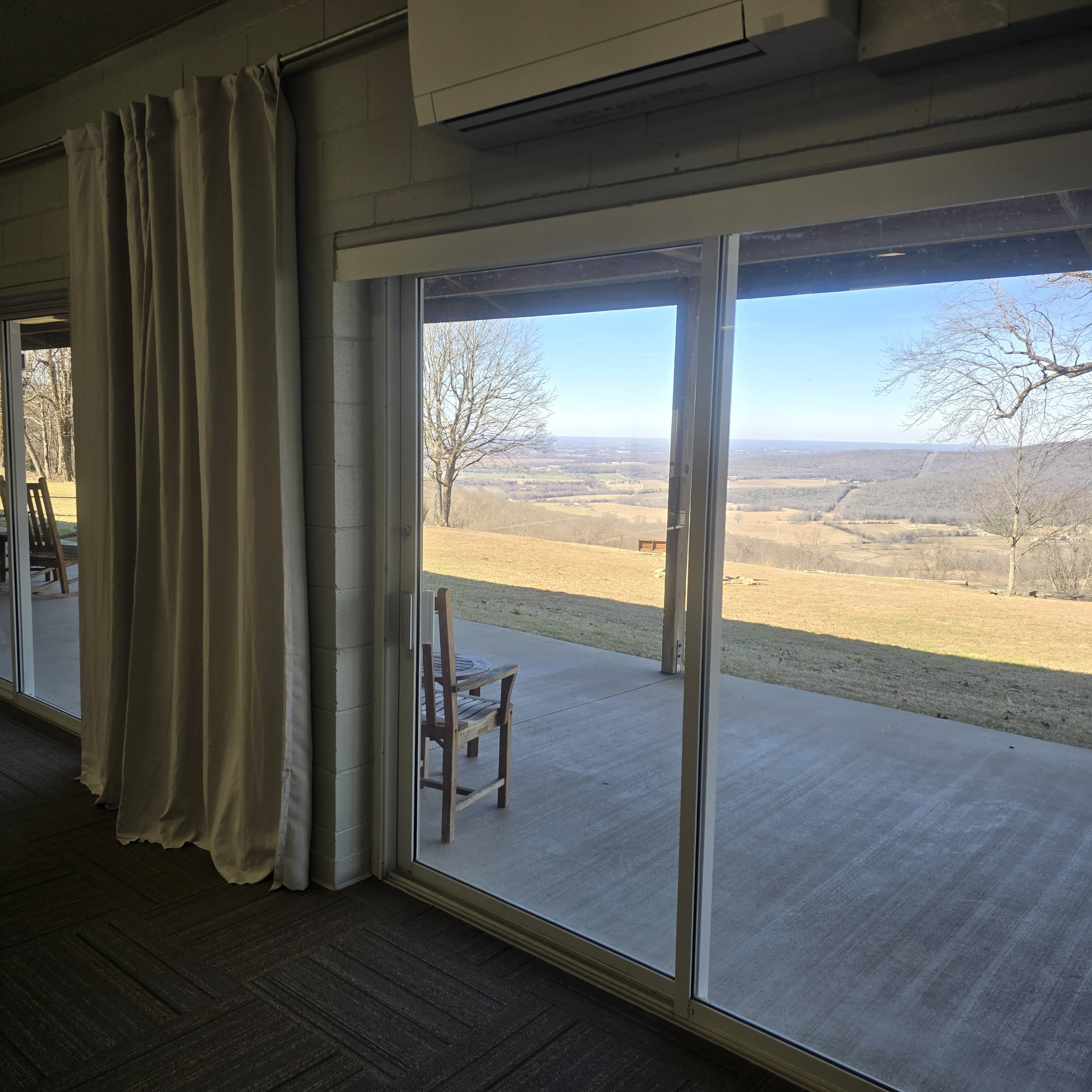 View of a hillside landscape with leafless trees and open fields from inside a room through sliding glass doors at a house.