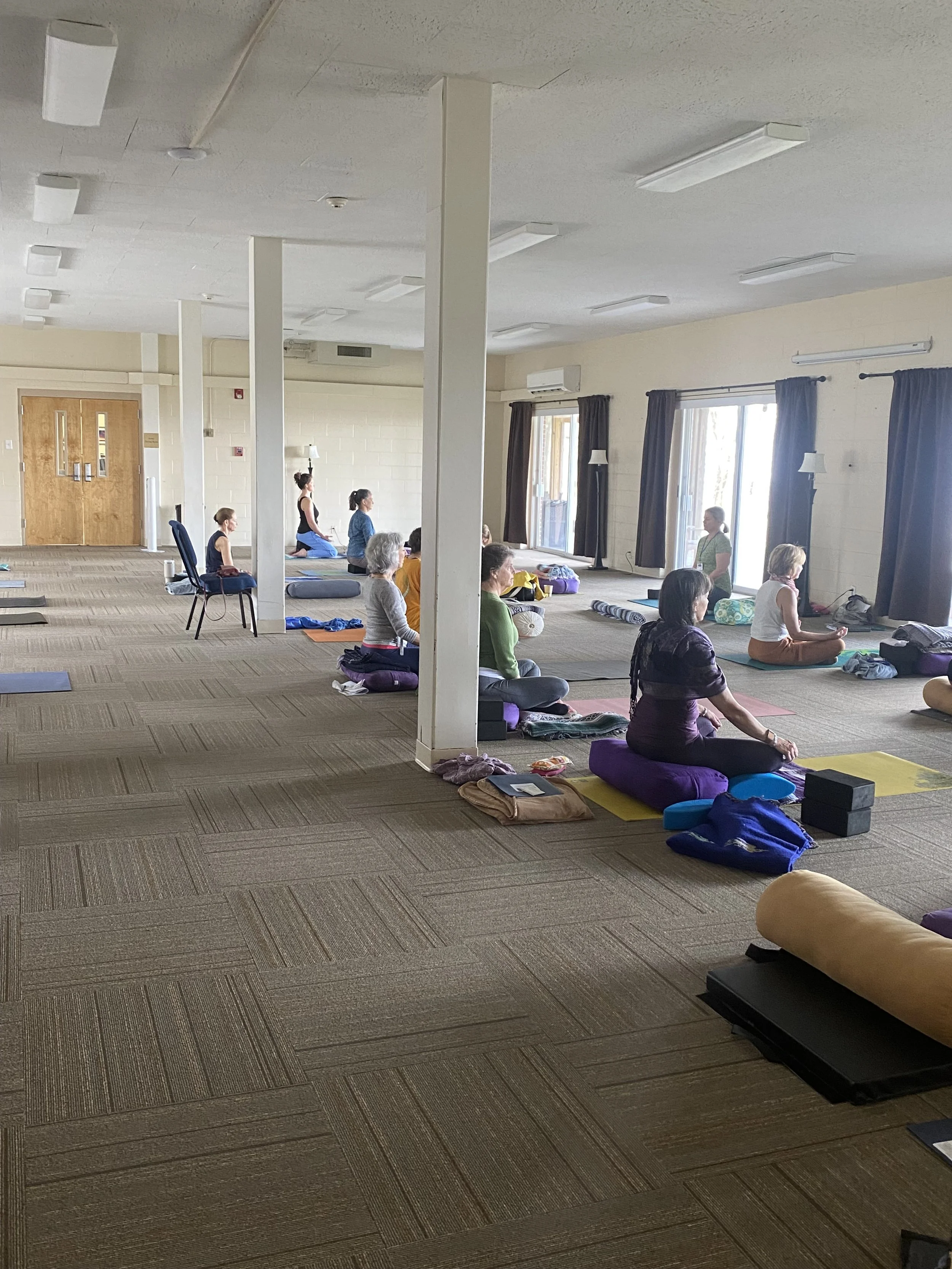 A group of women participating in a yoga class in a spacious, well-lit room with large windows and curtains, sitting on yoga mats.