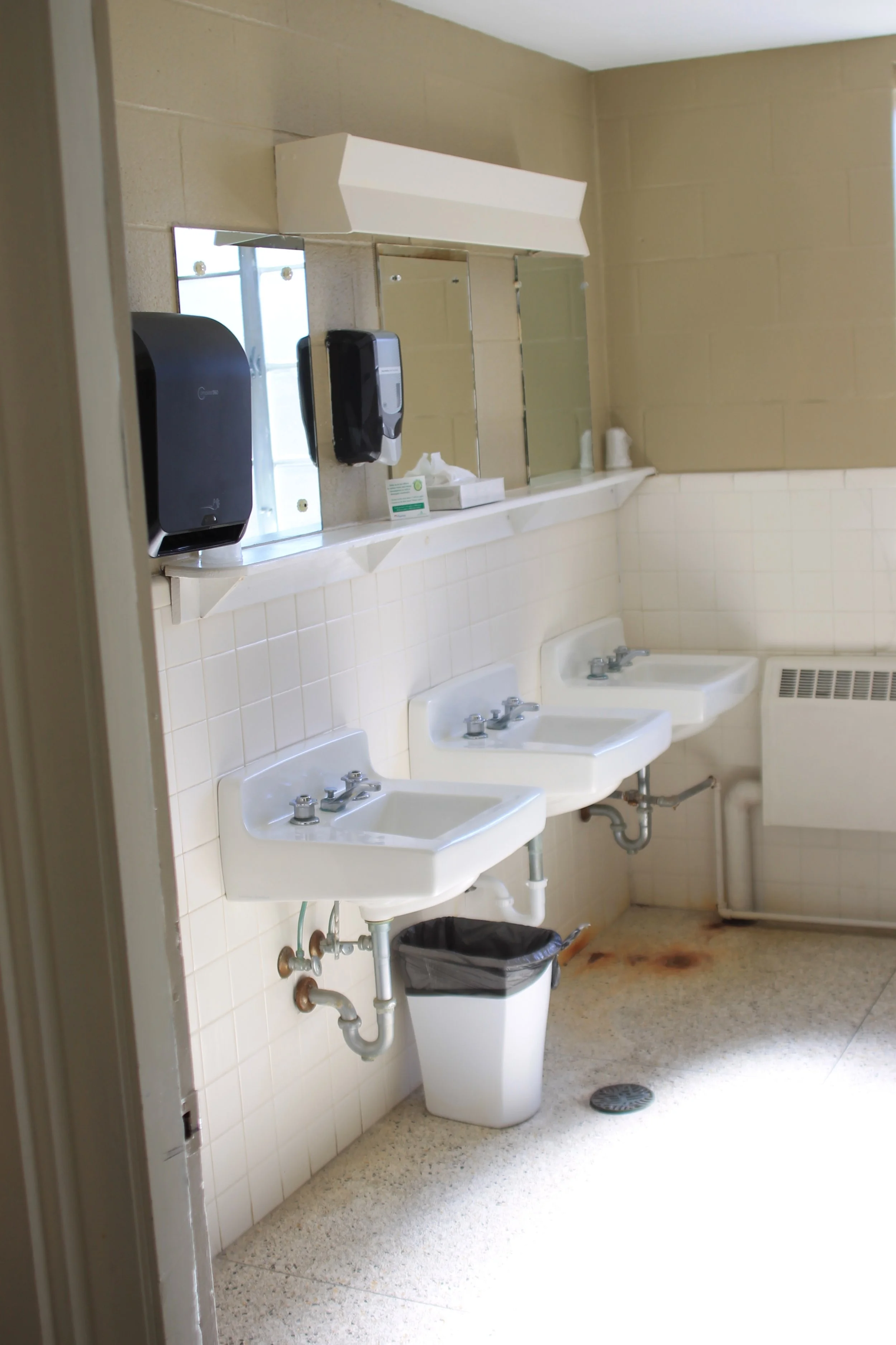 Three white sinks with silver faucets in a beige tiled bathroom with a trash can underneath and a vent on the wall.