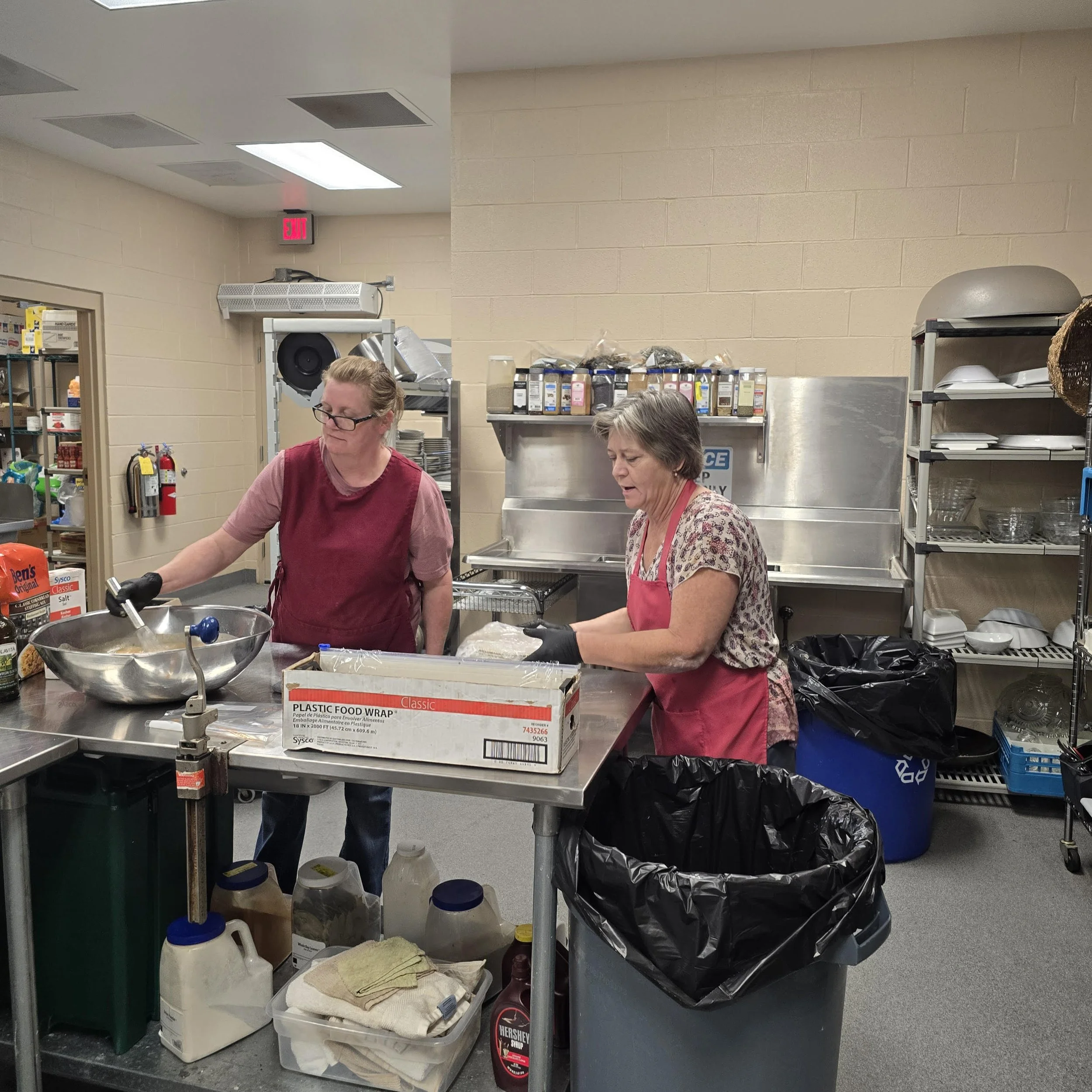 Two women preparing food in a commercial kitchen, one stirring a large metal bowl and the other wrapping food with plastic wrap.