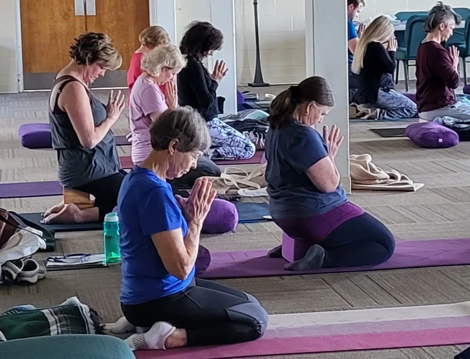 Group of people practicing yoga or meditation in a room, sitting on mats with their eyes closed and hands in prayer position.