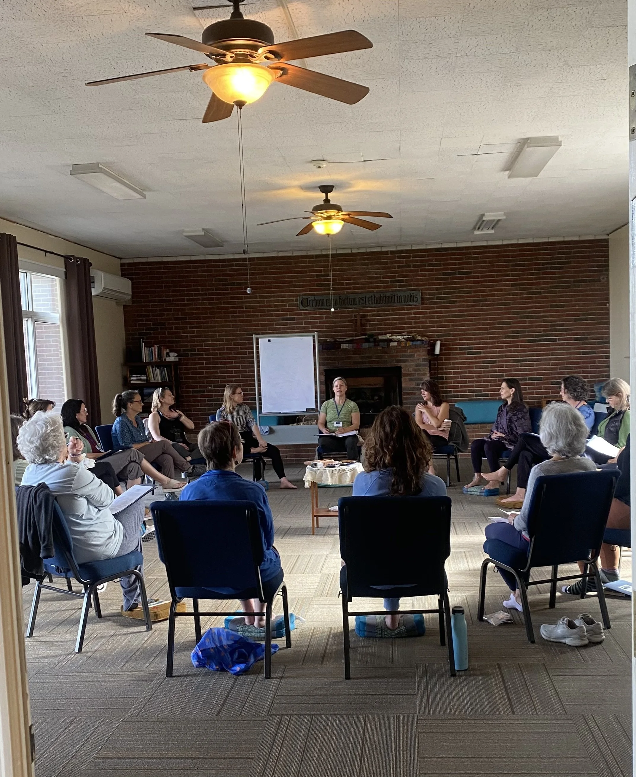 A group of people sitting in a circle in a room, participating in a workshop or meeting. The room has brick walls, ceiling fans, and windows with curtains.