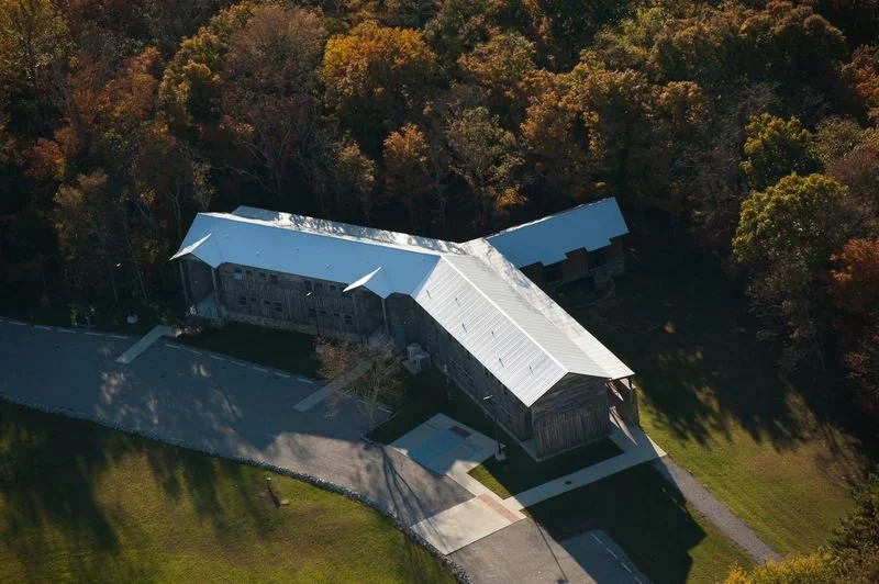 A large barn with a metal roof surrounded by trees and a paved driveway in autumn.