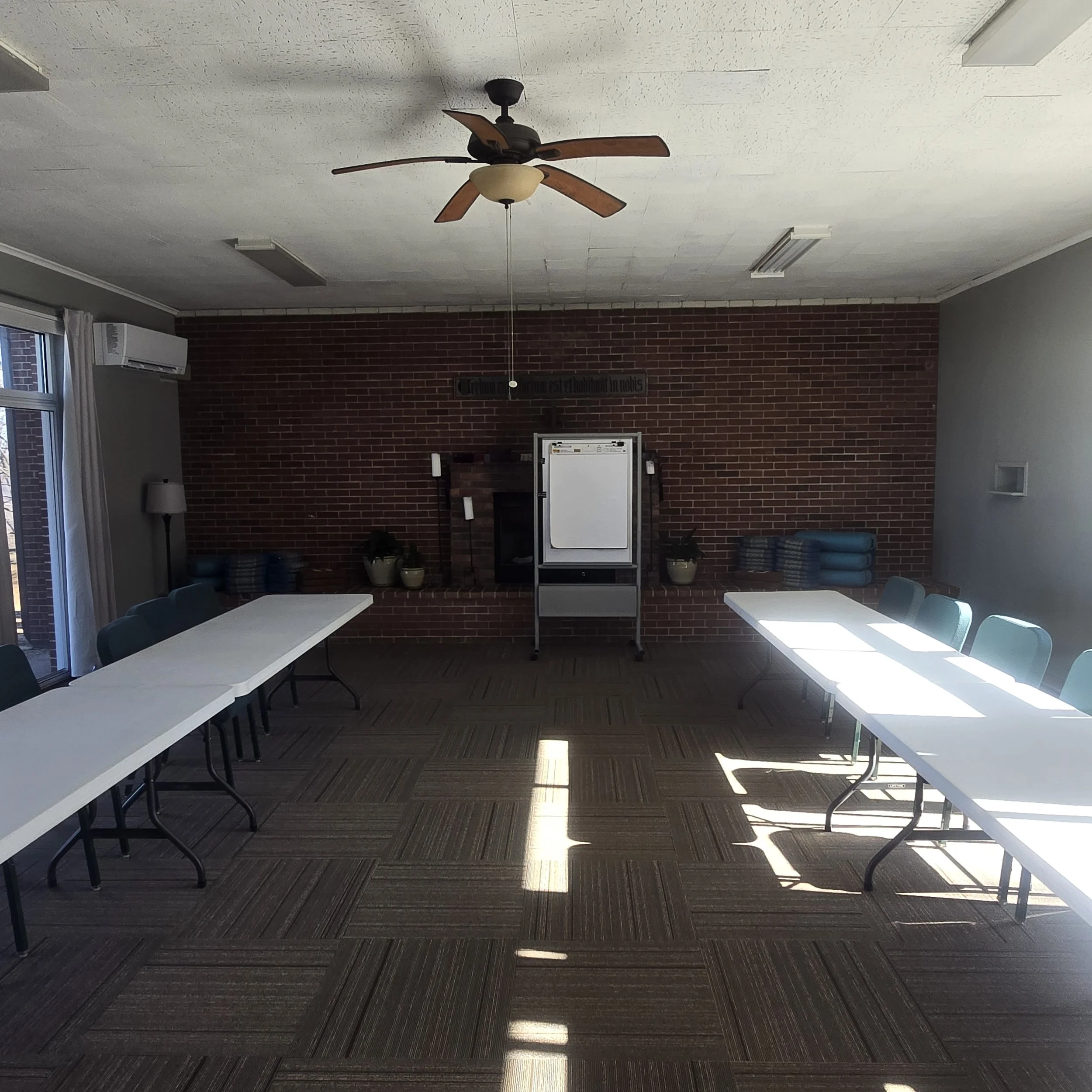 Meeting room with two long tables, green chairs, a brick fireplace, potted plants, folded blue blankets, a whiteboard, and windows with curtains, lit by natural light.