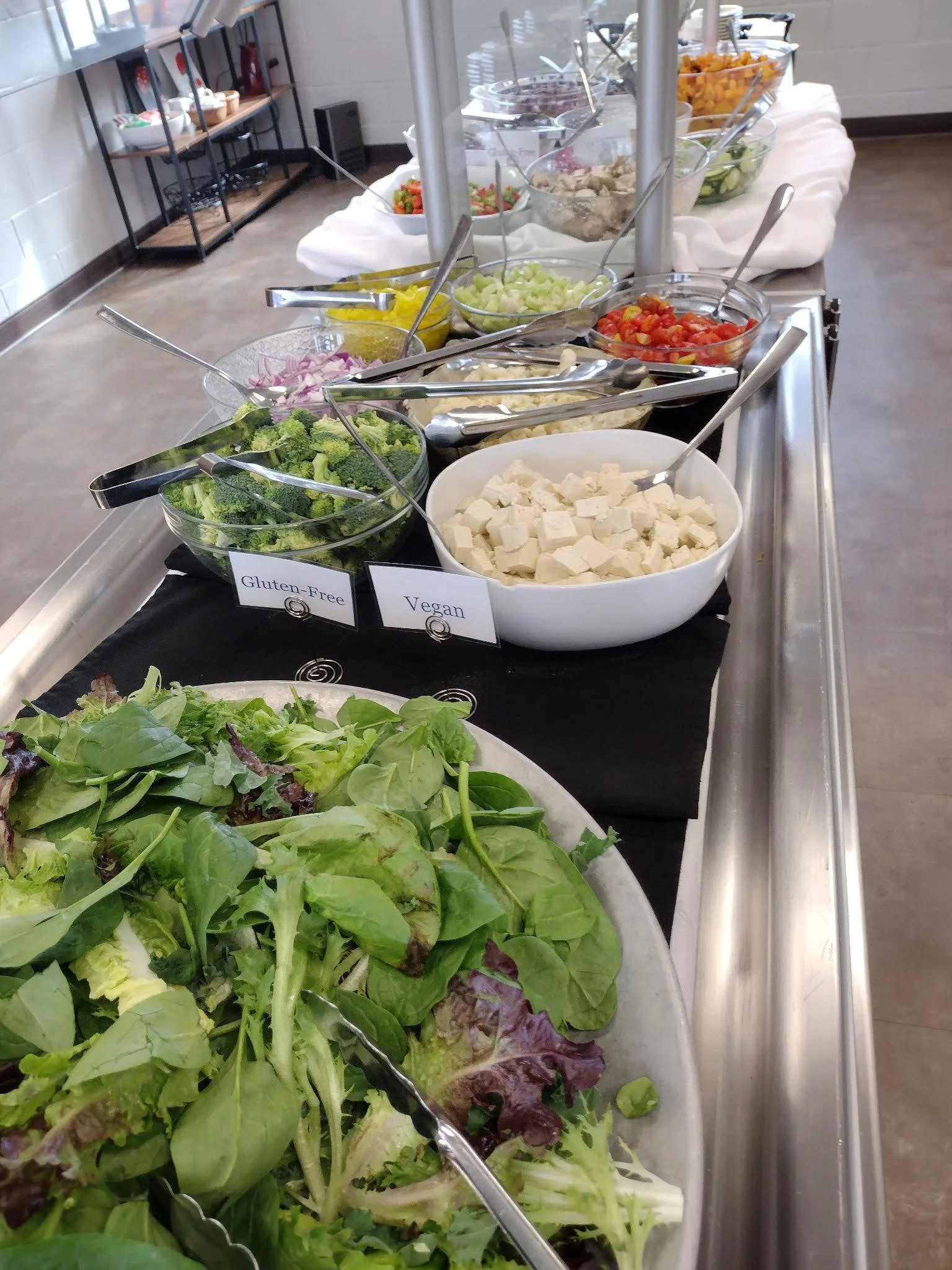 Buffet of fresh vegetables, including leafy greens, broccoli, cucumbers, tomatoes, chopped onions, coconut, and other salad toppings with labels indicating gluten-free and vegan options.