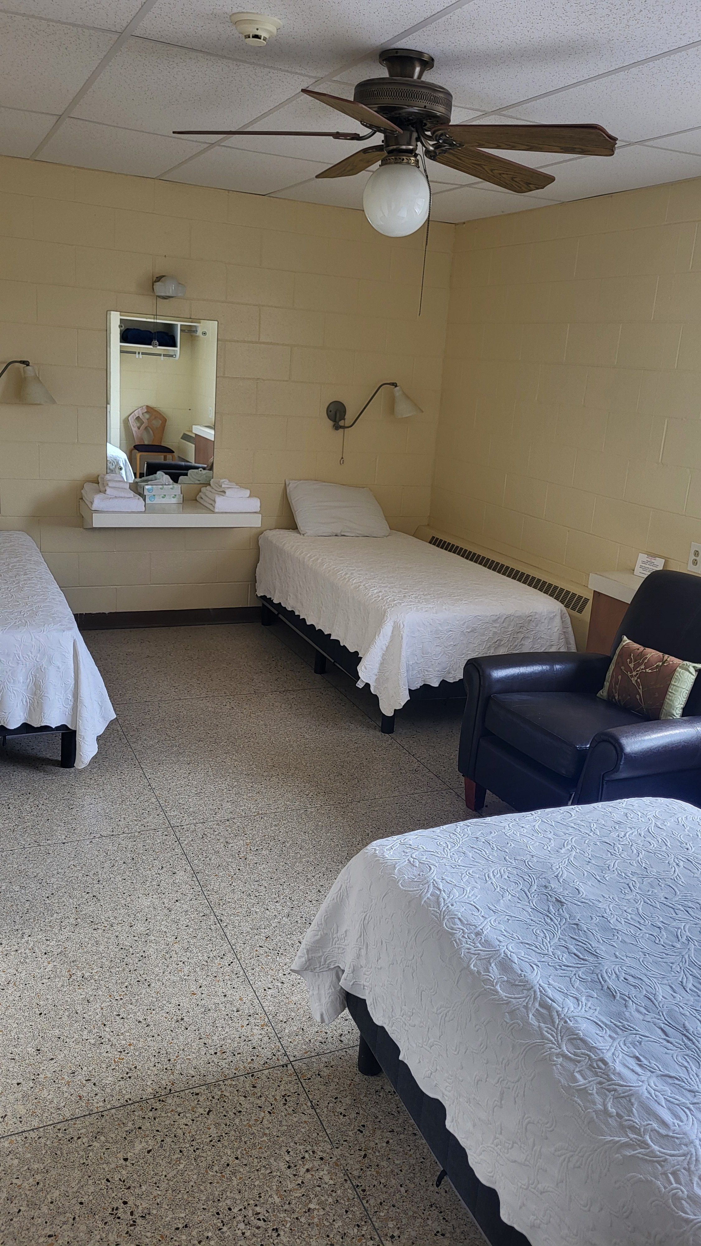 A hospital room with three beds covered in white, embroidered bedspreads. One bed is near the corner, another in the foreground, and the third is against the far wall, with a pillow and a headboard. There are two wall-mounted lamps above two beds, a 