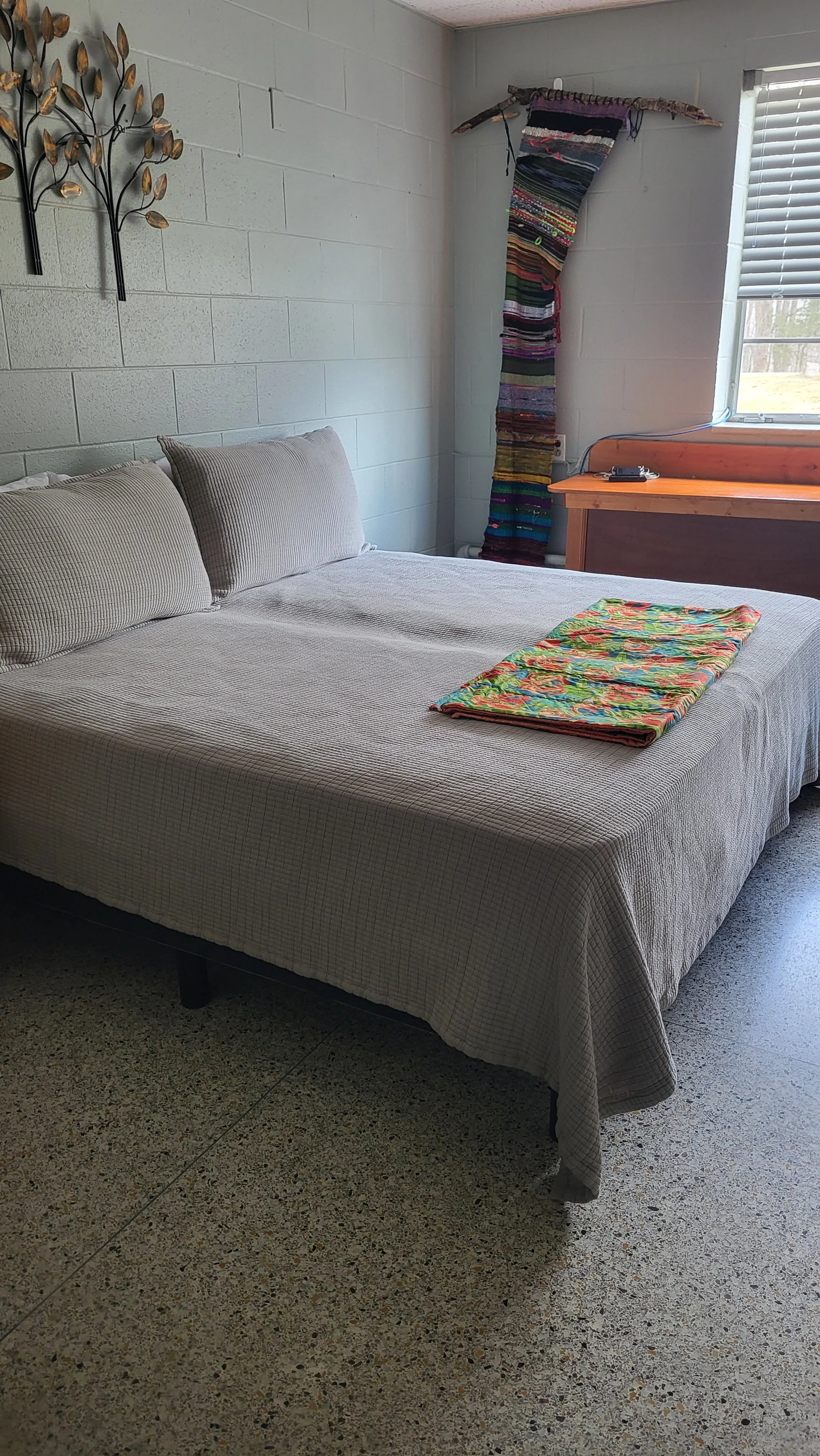 A bedroom with a bed covered in a beige checked bedspread, two pillows, a colorful fabric on the bed, a wall decoration of metal leaves, a fabric wall hanging, a wooden desk with a phone charger, and a window with blinds.