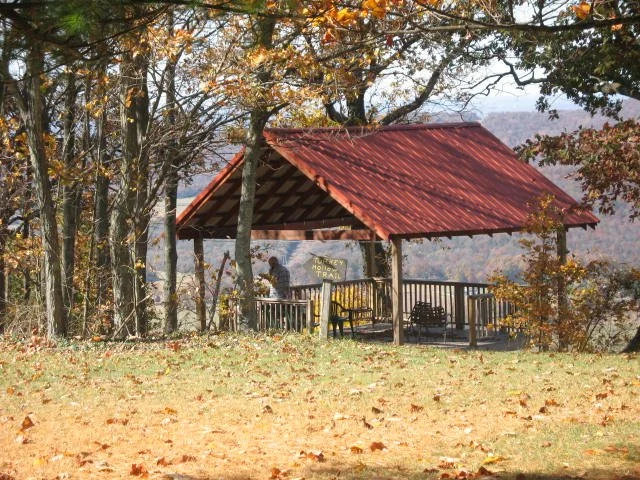 A scenic overlook shelter with a red roof and wooden railings, surrounded by fall trees, overlooking a mountain valley.