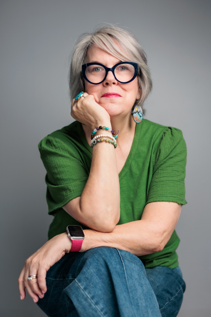 A woman with gray hair and glasses holding an open book in a bookstore or library.