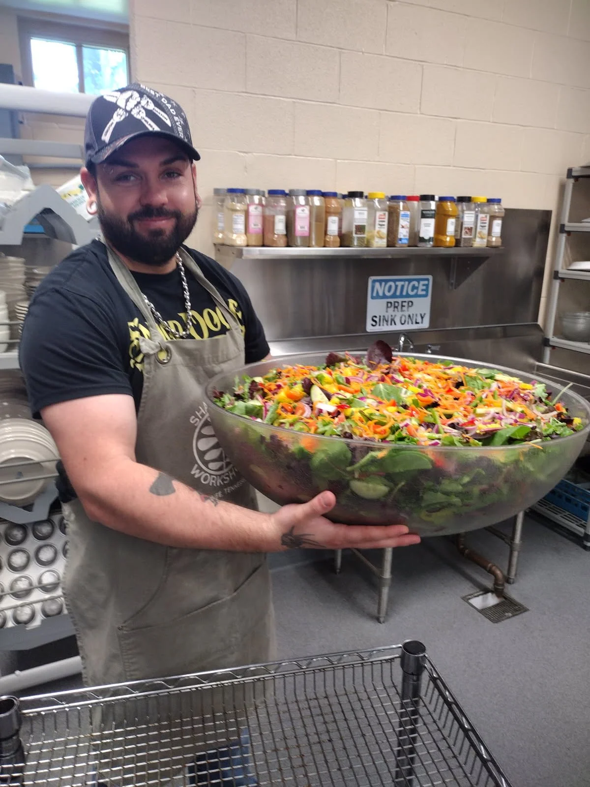 A man with a beard, wearing a black t-shirt, gray apron, and a black cap, is holding a large bowl of colorful salad in a kitchen.