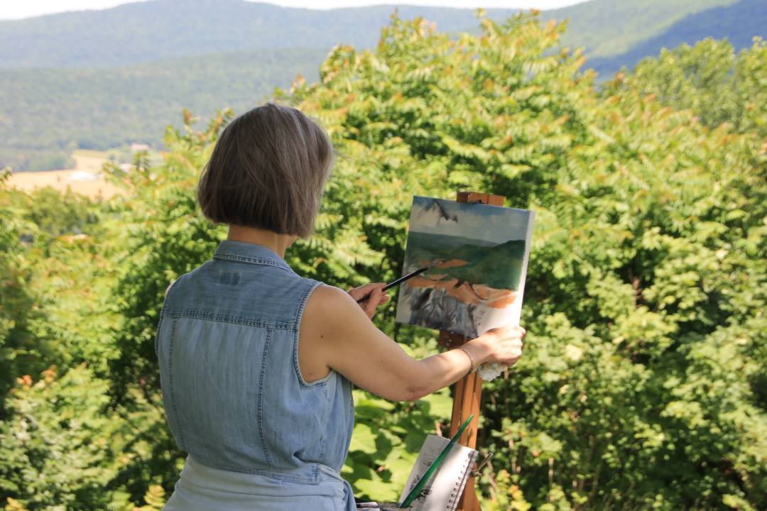 A woman with short brown hair in a sleeveless denim vest, painting a landscape on an easel outdoors surrounded by green trees and distant mountains.